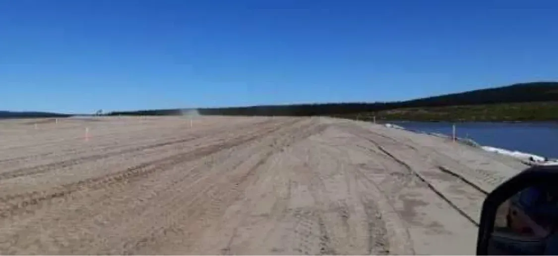 Un large chemin sablonneux ou une zone de construction s'étend vers un plan d'eau sous un ciel bleu limpide.
