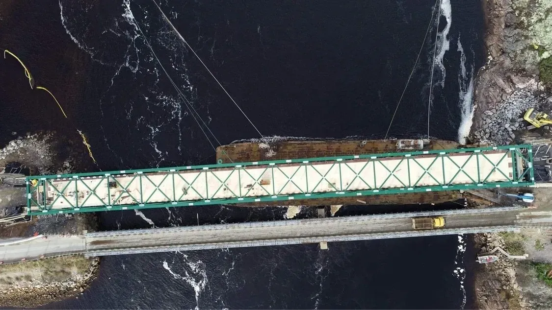 Vue aérienne d'un pont à treillis vert enjambant une rivière sombre, à côté d'une route étroite traversée par un véhicule.