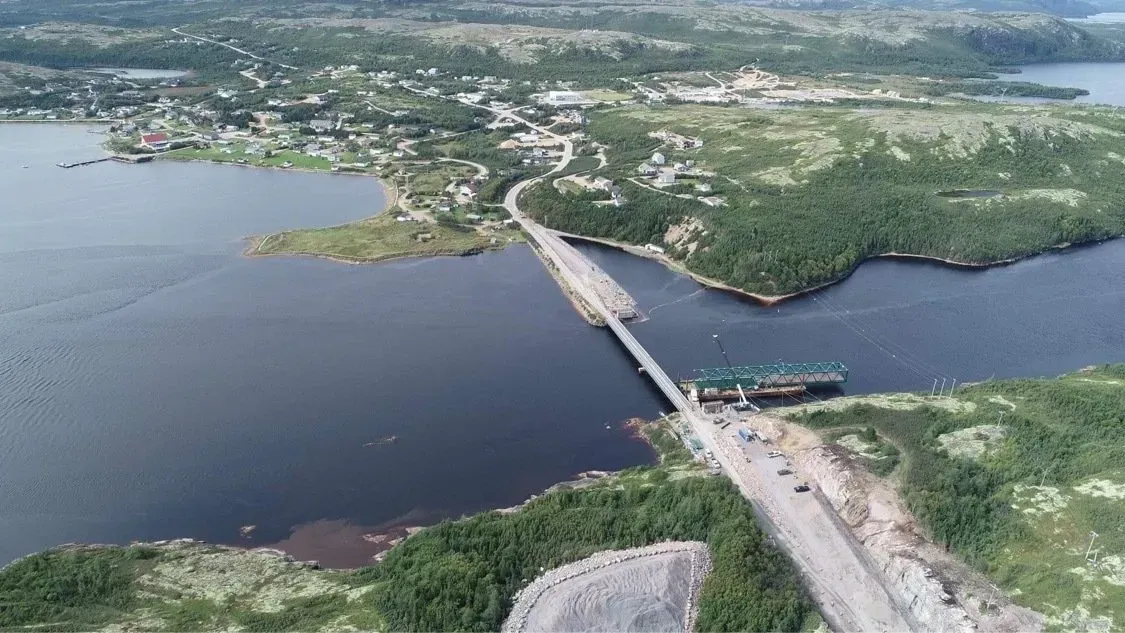 Vue aérienne d'un pont reliant les deux rives d'une étendue d'eau près d'un village côtier et d'un paysage rocheux verdoyant.