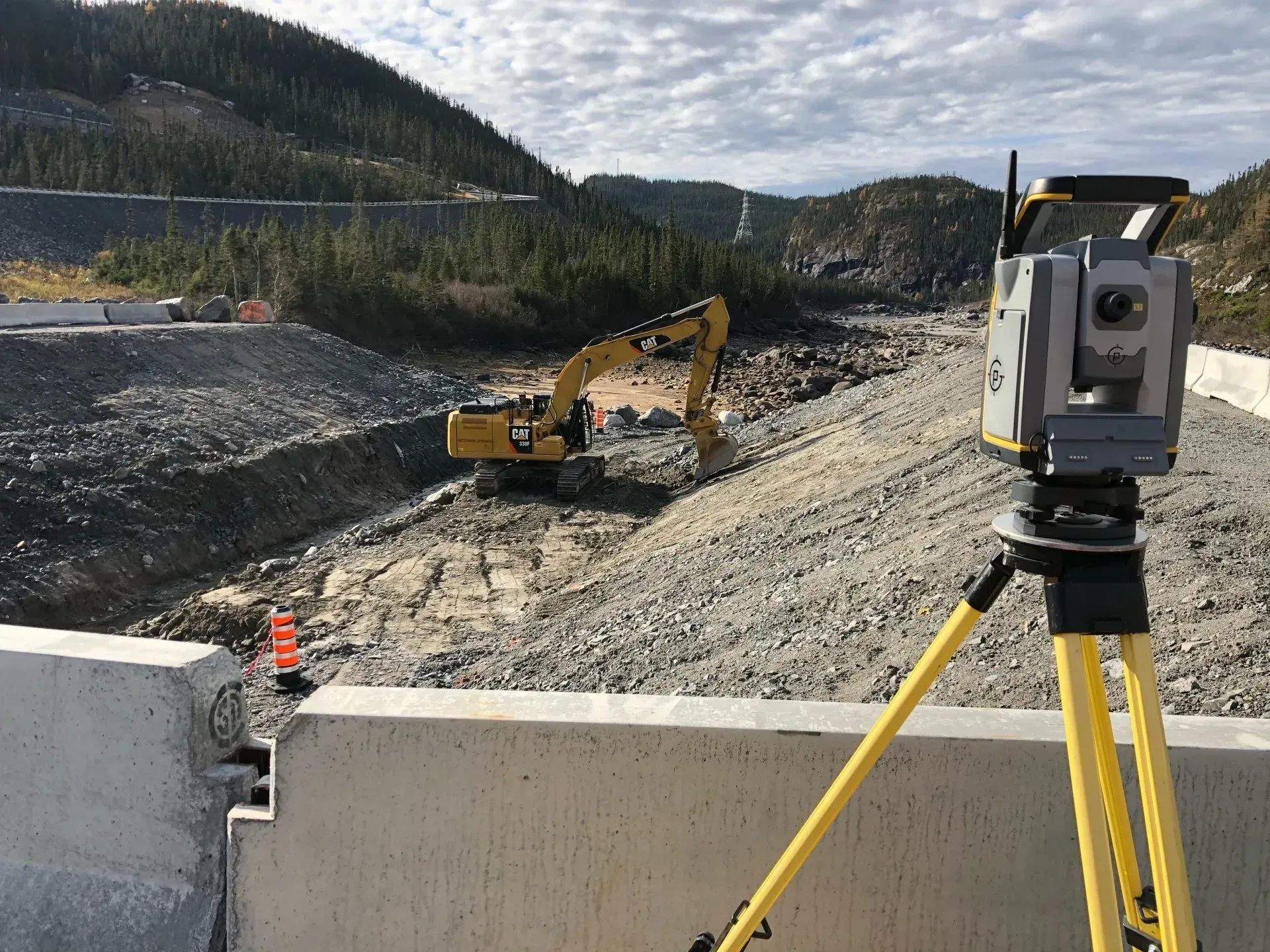 Une station totale de topographie, montée sur un trépied, surplombe une excavatrice jaune en activité sur un chantier rocailleux.