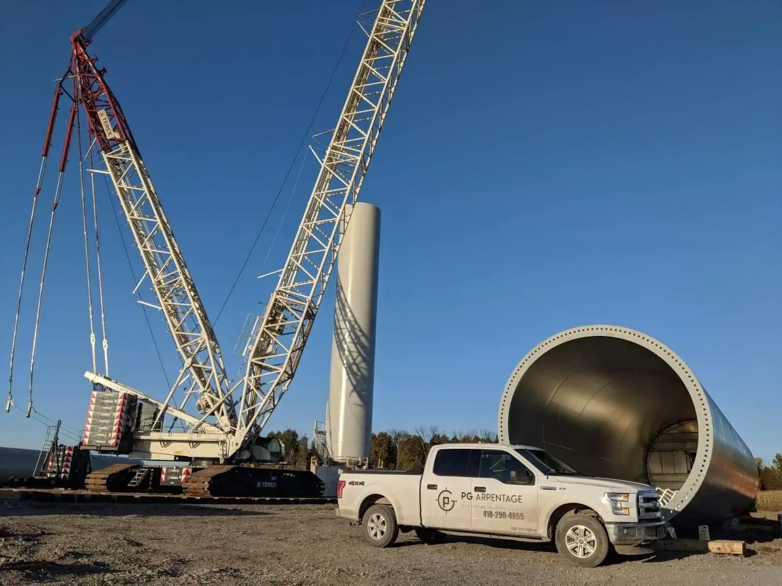 Grue et ouvriers près d'une immense structure cylindrique blanche sur un chantier de construction en plein air, sous un ciel bleu dégagé.