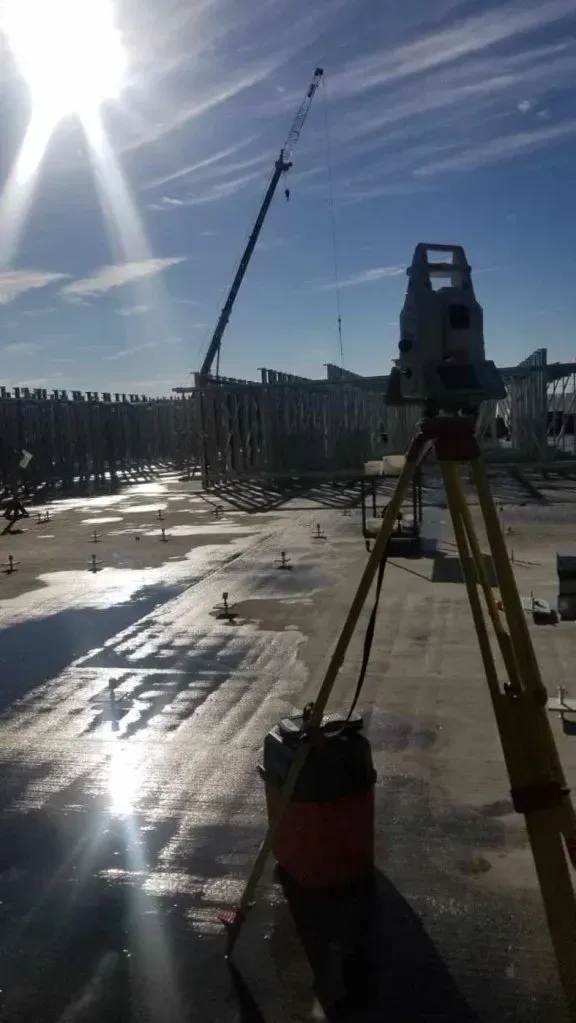 Une station totale de topographie, installée sur un trépied, surplombe un chantier ensoleillé où se dresse une grue au loin.