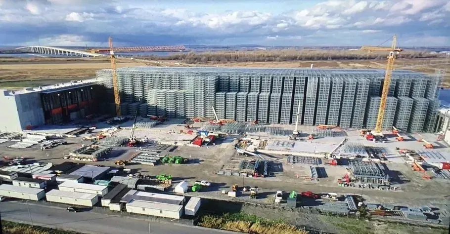 Vue aérienne d'un grand chantier de construction d'entrepôt, caractérisé par une longue façade en grille métallique et deux grues imposantes.