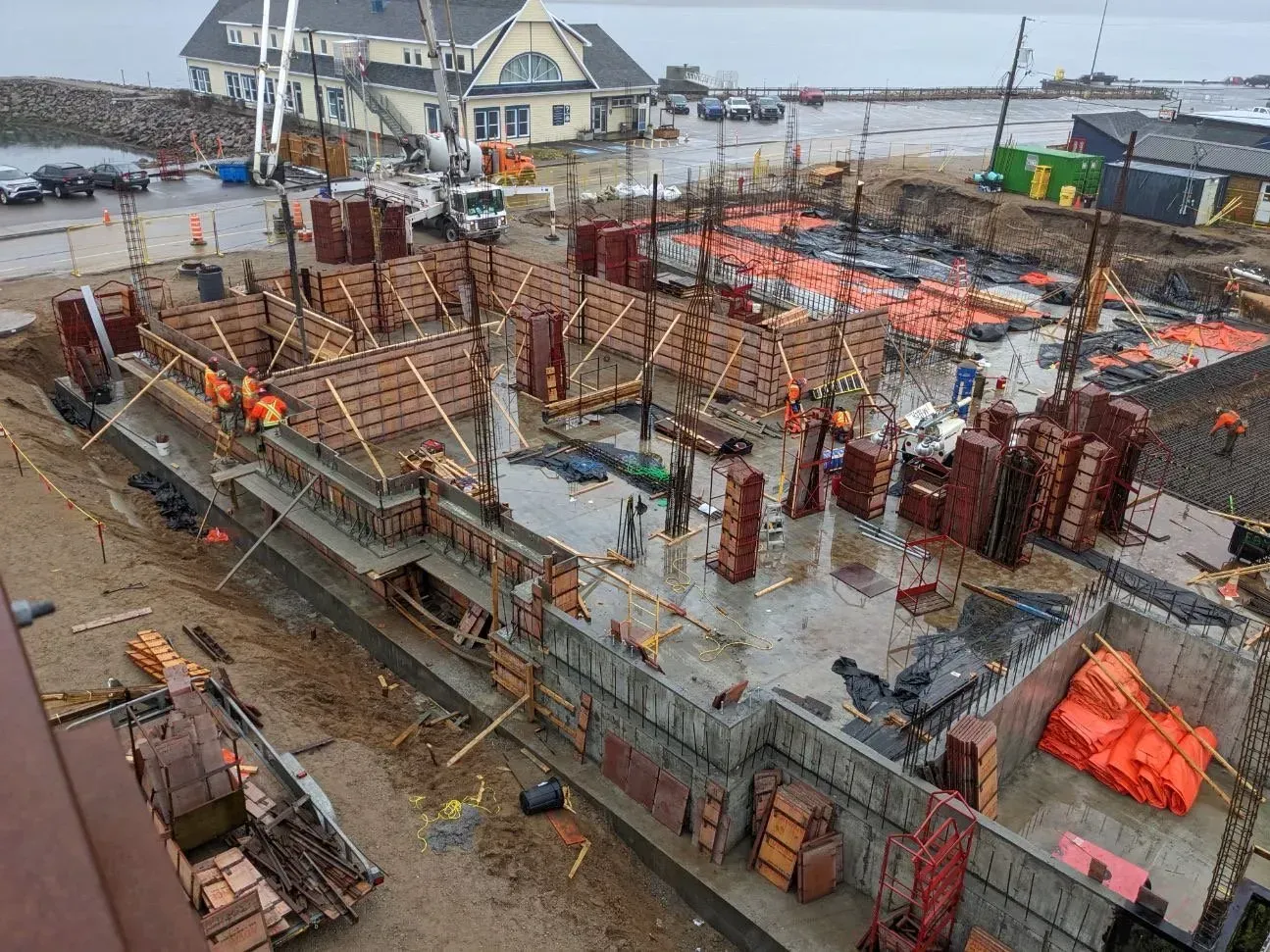 Chantier de construction avec fondations en béton, armatures en acier, ouvriers et barrières de sécurité orange près de l'eau