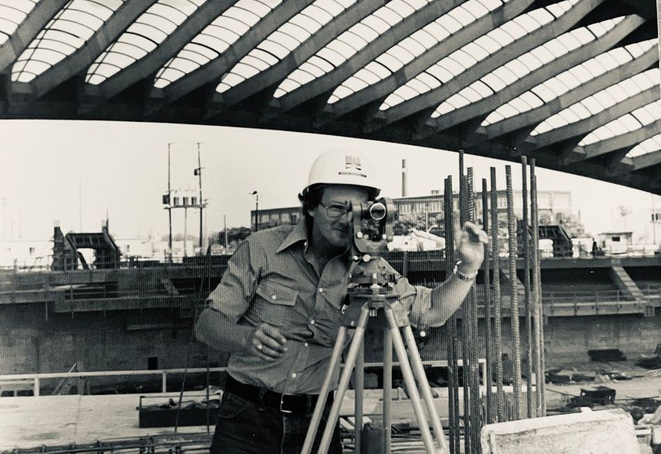 Un géomètre portant un casque de chantier observe un chantier de construction doté d'un grand toit en béton nervuré à travers un théodolite placé sur un trépied.