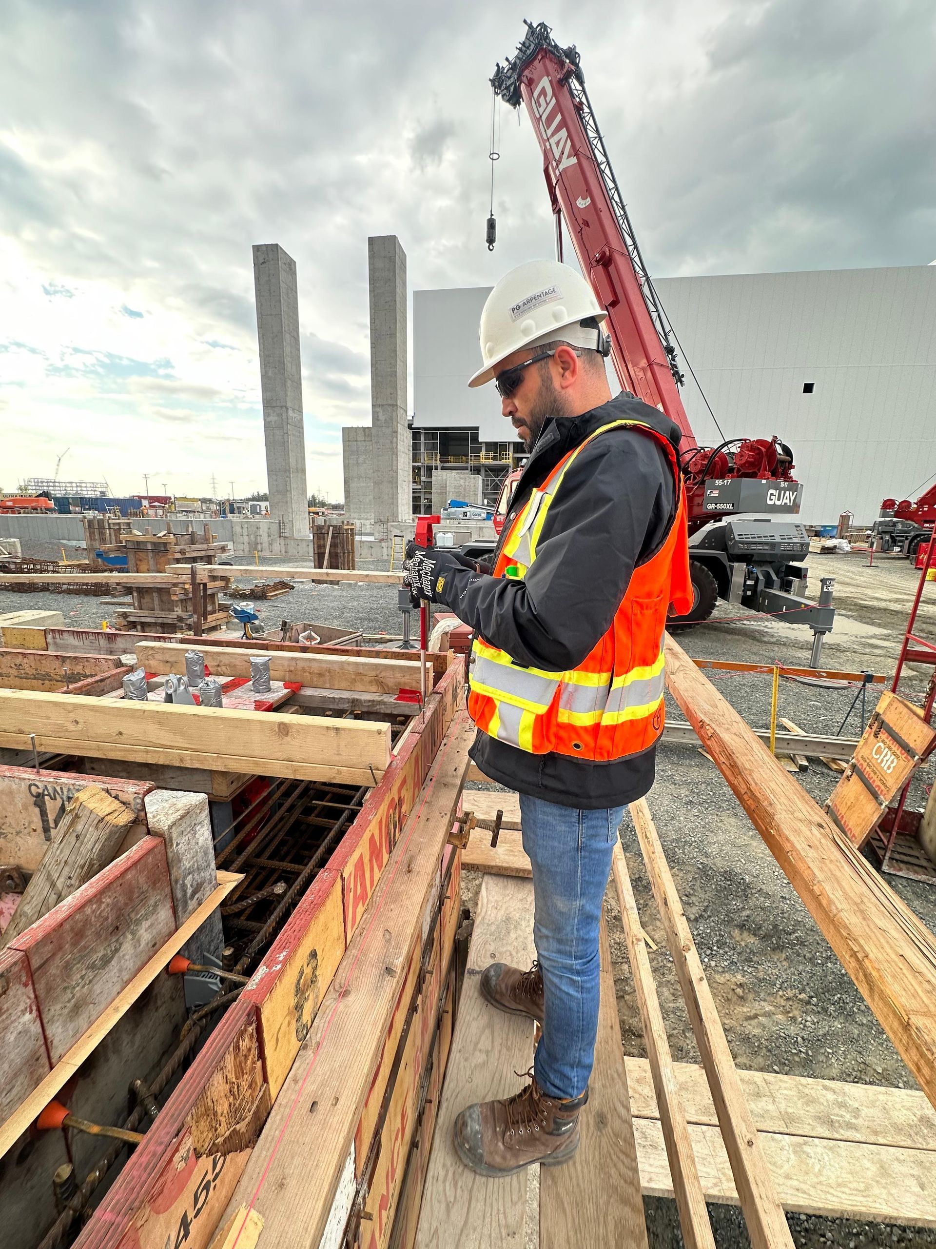 Un ouvrier du bâtiment, casqué et vêtu d'un gilet de sécurité, se tient sur un chantier à côté de barres d'armature et d'une grue.