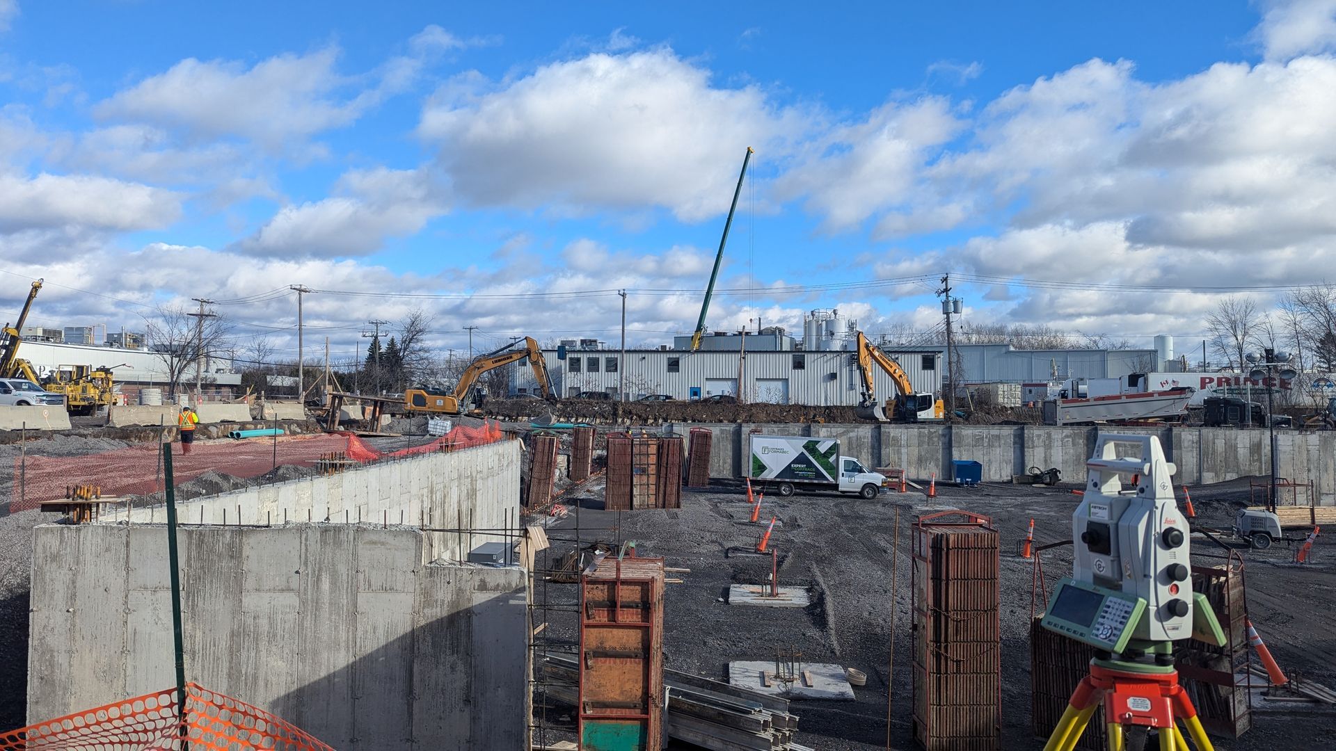 Chantier de construction avec murs en béton, grues et ouvriers sous un ciel bleu nuageux.