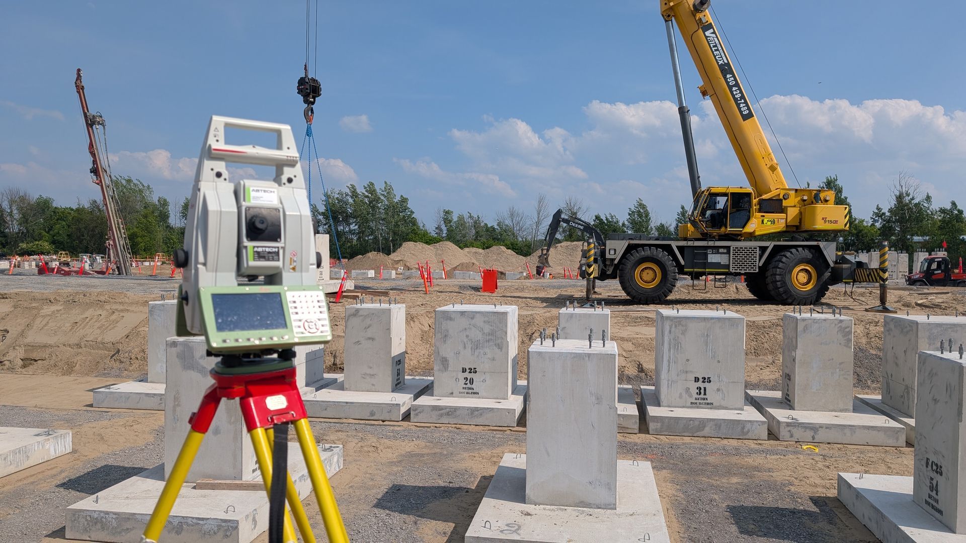 Une station totale de topographie, montée sur un trépied jaune, surplombe un chantier de construction avec des piliers en béton et une grue mobile jaune.
