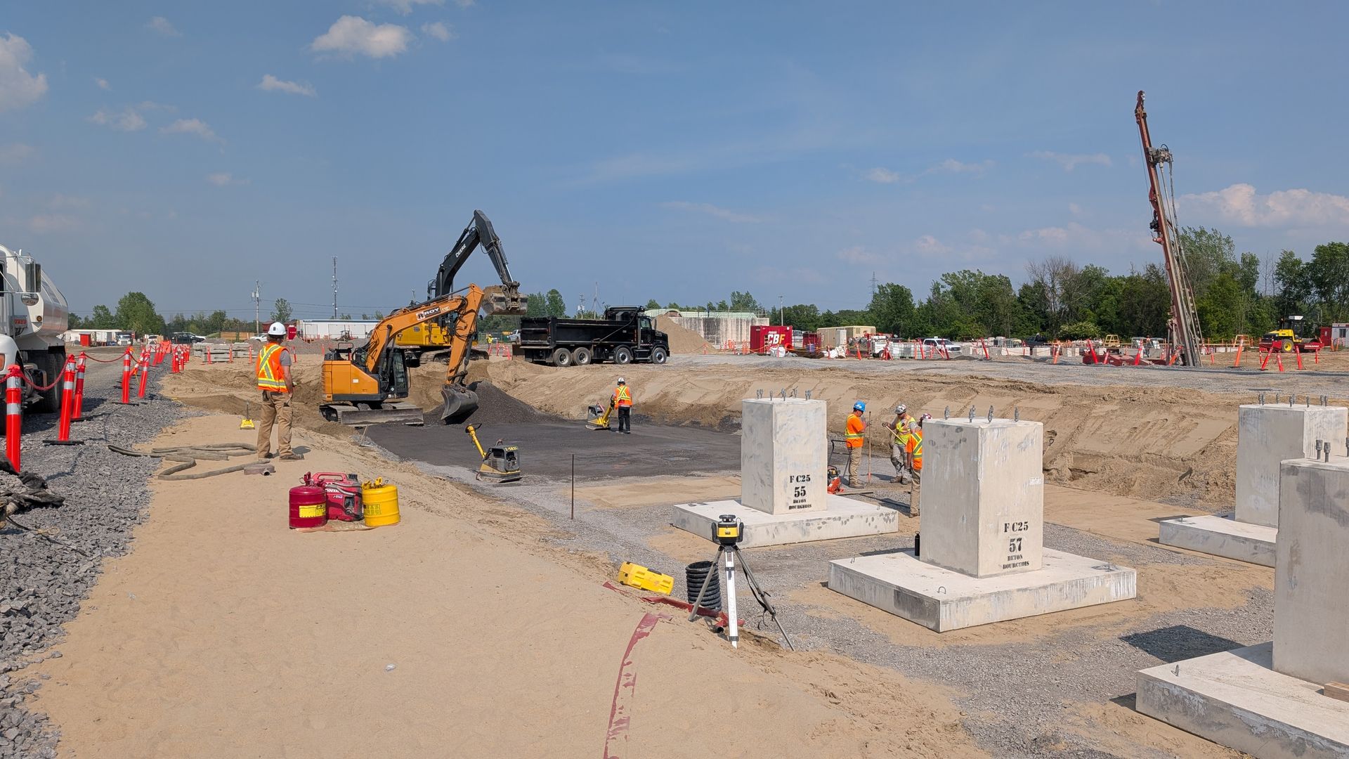 Des ouvriers du bâtiment construisent des piliers en béton sur un chantier ensoleillé à l'aide d'engins lourds et de gravier.