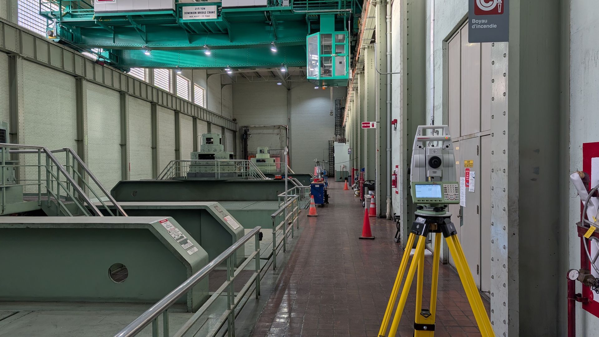 Station totale sur trépied jaune dans un bâtiment industriel avec équipements verts et panneaux de sécurité.