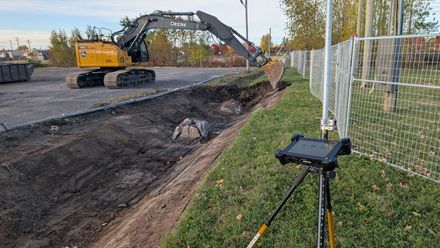 Un chantier de construction avec une pelleteuse jaune creusant une tranchée, vu depuis une tablette posée sur un trépied.
