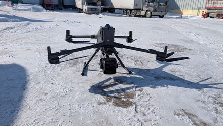 Un drone industriel noir est posé sur un sol enneigé, avec des camions stationnés en arrière-plan.