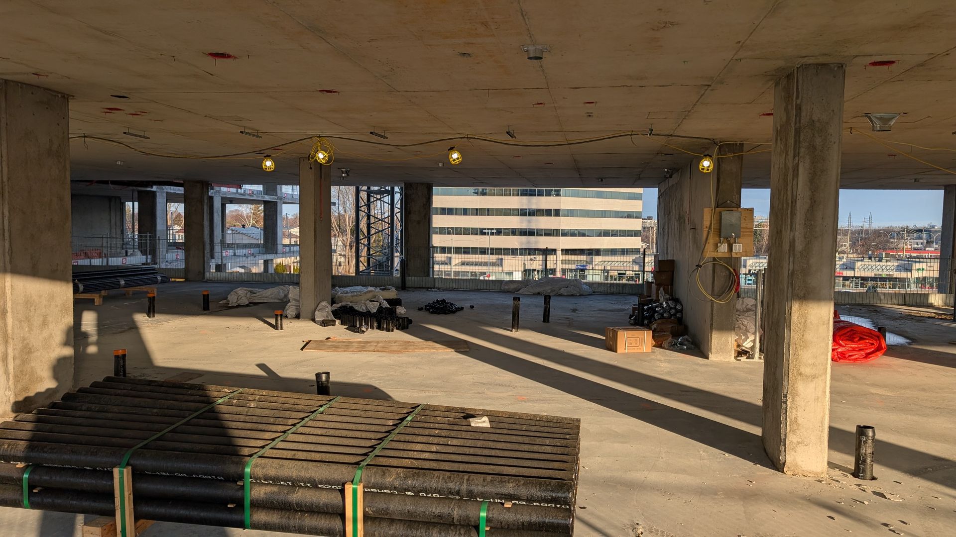 Intérieur d'un chantier de construction vide, avec des piliers en béton, des matériaux empilés et la lumière du soleil qui filtre à travers les ouvertures.