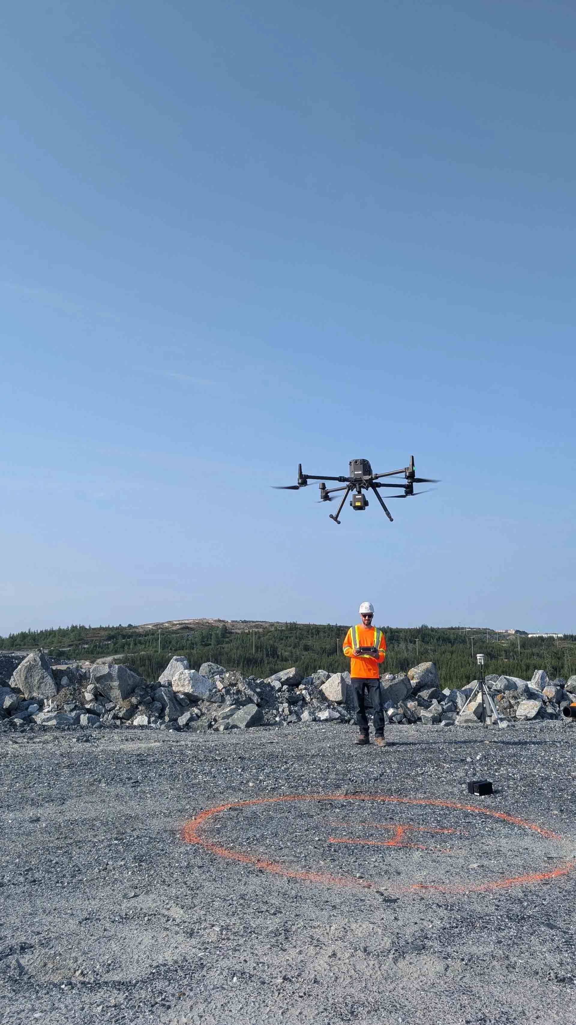Un drone survole une personne portant un gilet orange sur un champ rocailleux sous un ciel bleu dégagé.