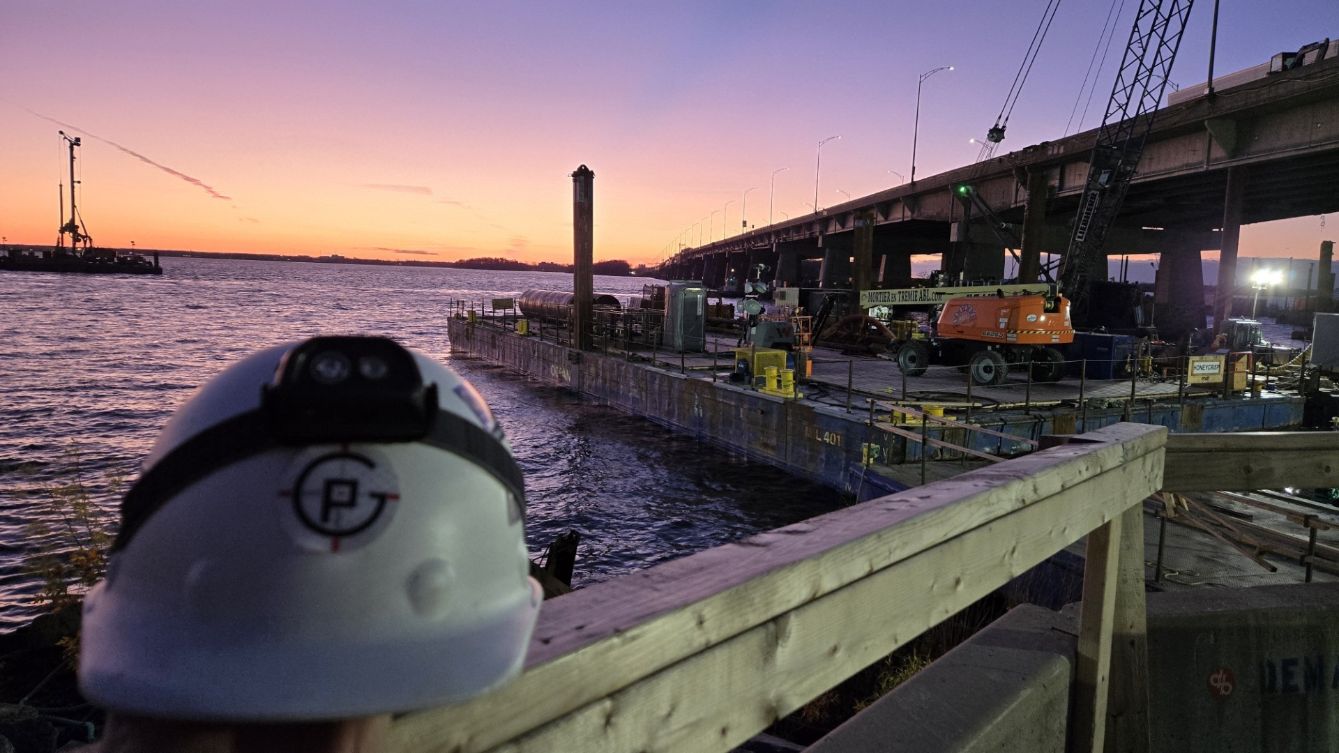 Casque de chantier au premier plan sur un chantier riverain sous un pont au coucher du soleil.