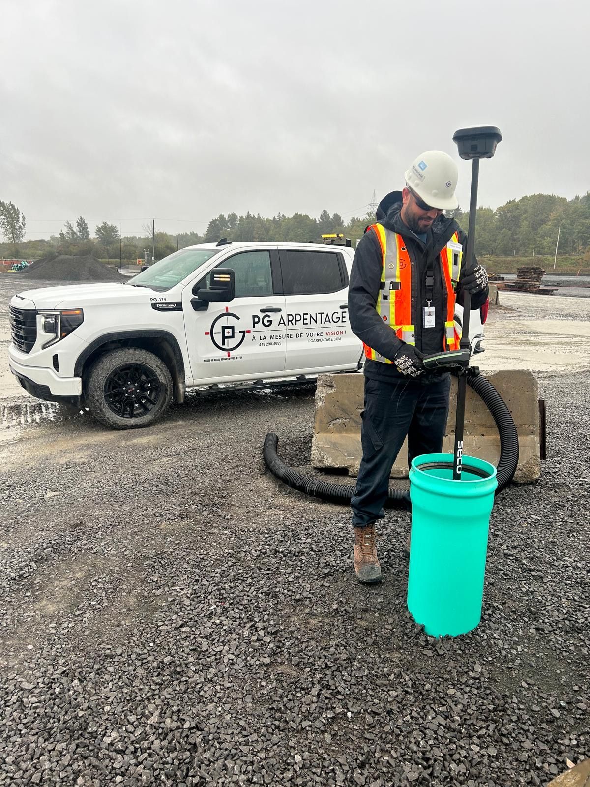 Un ouvrier portant un casque de chantier utilise un tuyau d'arrosage à côté d'une camionnette blanche sur un chantier de gravier.