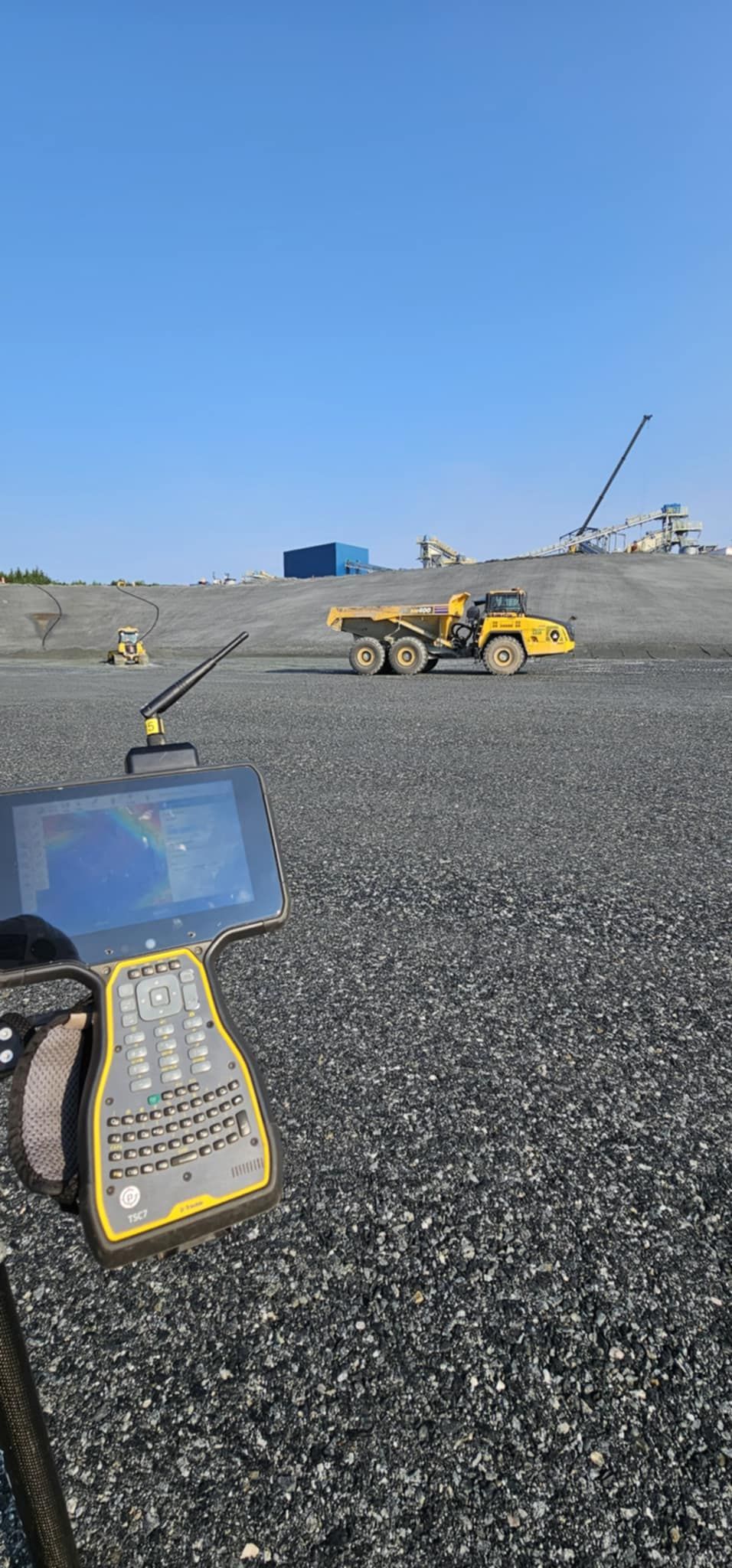 Matériel topographique sur un site de carrière rocailleux, avec une chargeuse jaune et des tas de gravier sous un ciel bleu.