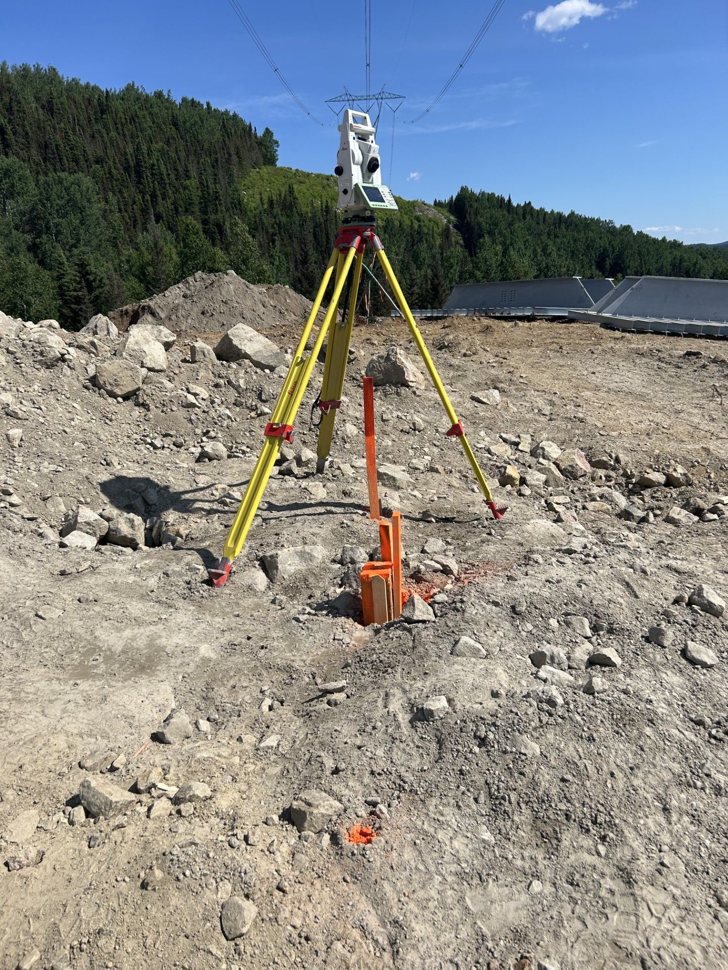 Trépied de topographie sur un chantier rocailleux sous un ciel bleu, avec des collines et des arbres en arrière-plan
