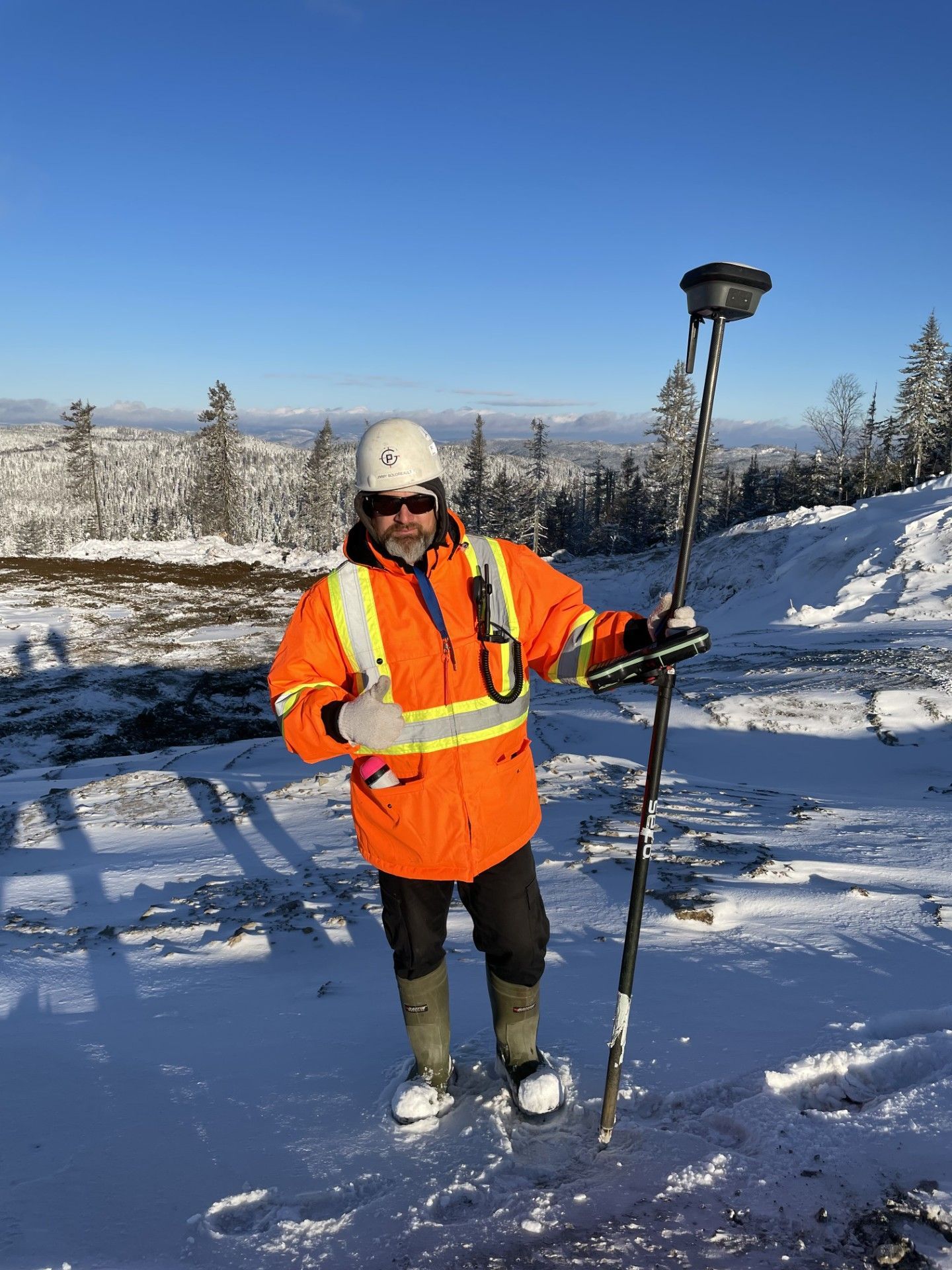 Une personne portant une veste de sécurité orange et un casque se tient debout dans un paysage enneigé, tenant un bâton de ski sous un ciel bleu clair.