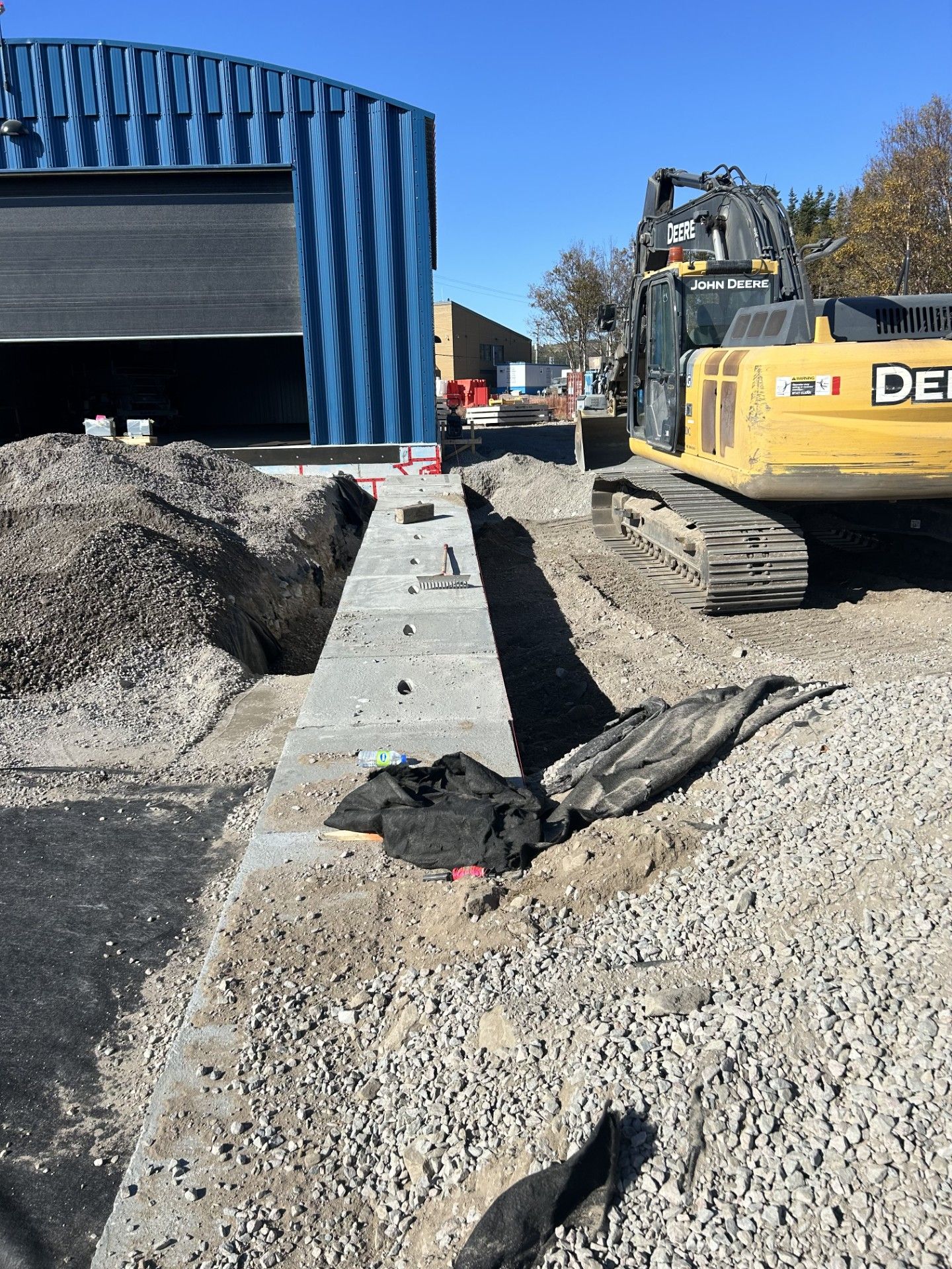 Chantier de construction avec une excavatrice à côté d'une tranchée en béton menant à un bâtiment industriel bleu.
