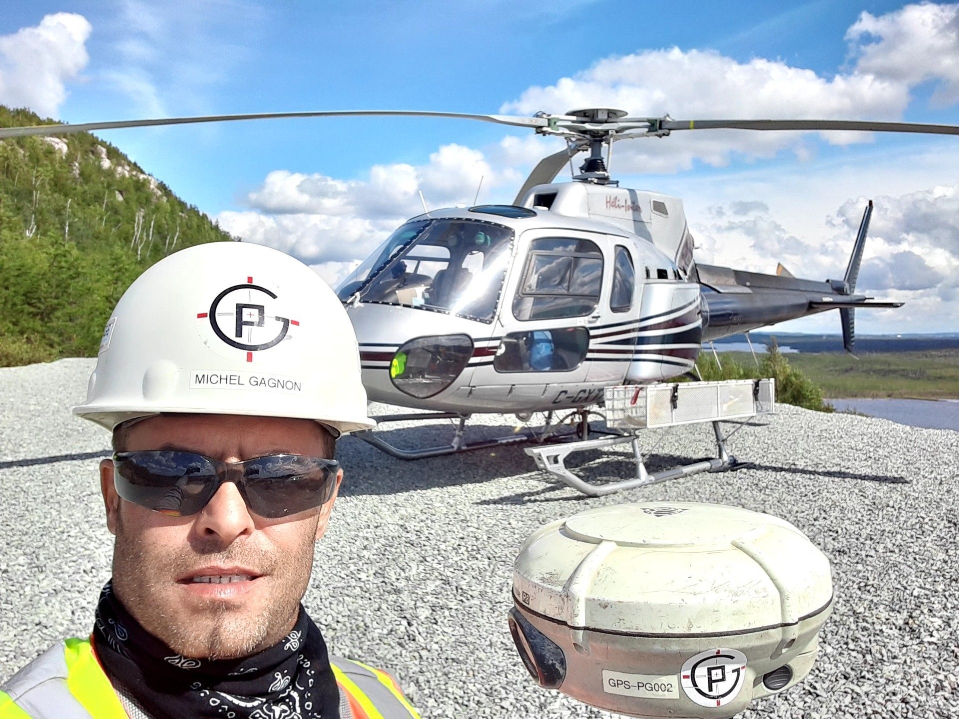 Hélicoptère sur une aire de gravier, avec un ouvrier portant un casque au premier plan et un lac de montagne en arrière-plan.