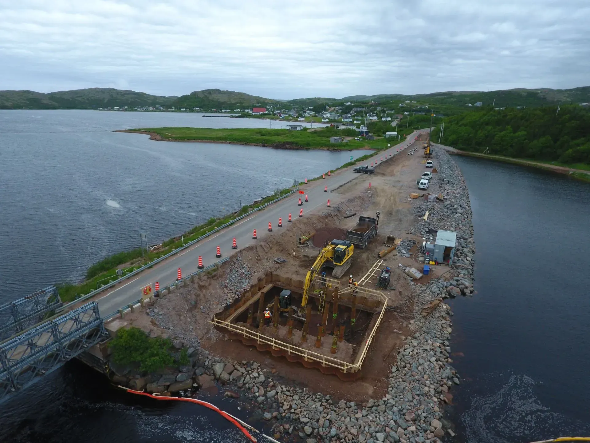 Vue aérienne d'une route côtière en construction, montrant une excavatrice travaillant dans une fosse rectangulaire près d'un pont.