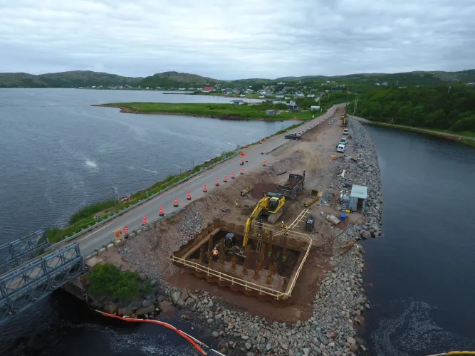 Vue aérienne d'une route côtière en construction, montrant une excavatrice travaillant dans une fosse rectangulaire près d'un pont.