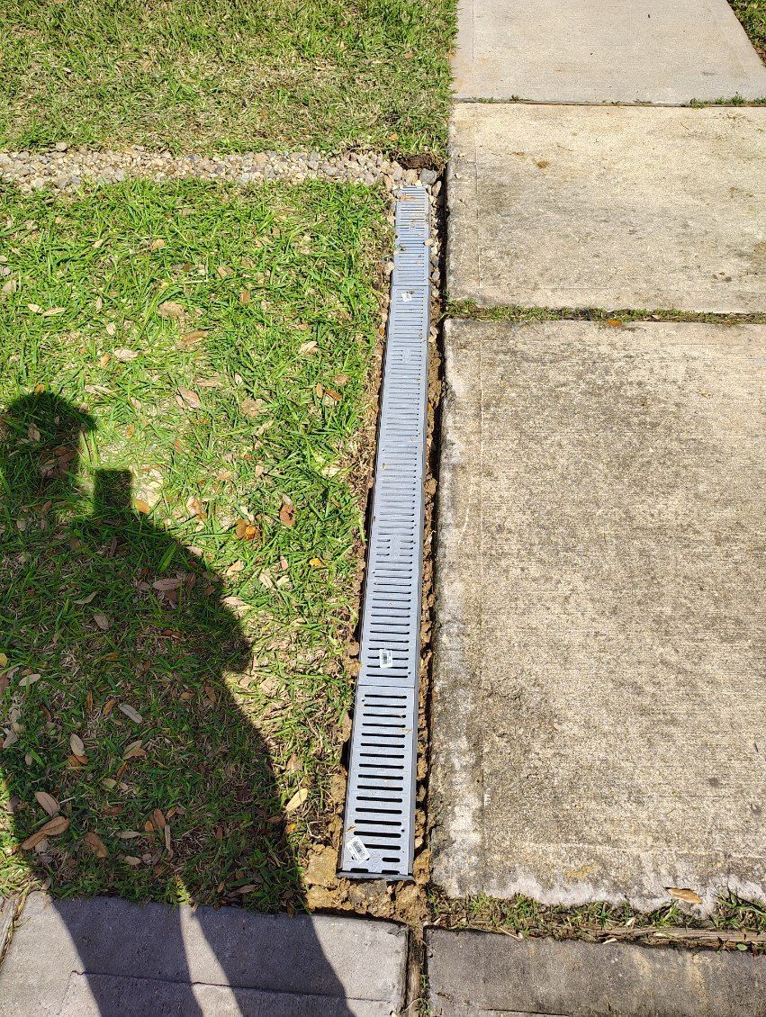 A long, metal drain runs alongside a sidewalk, between the grass and concrete.