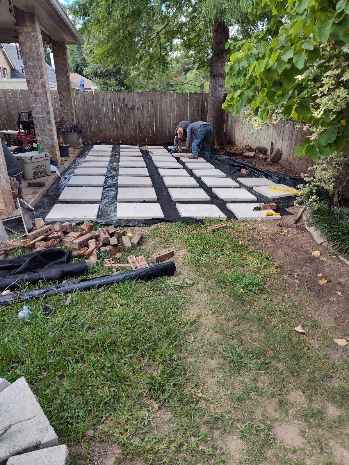 Backyard patio under construction with square pavers being laid out. Two people are working, grass and a fence are visible.