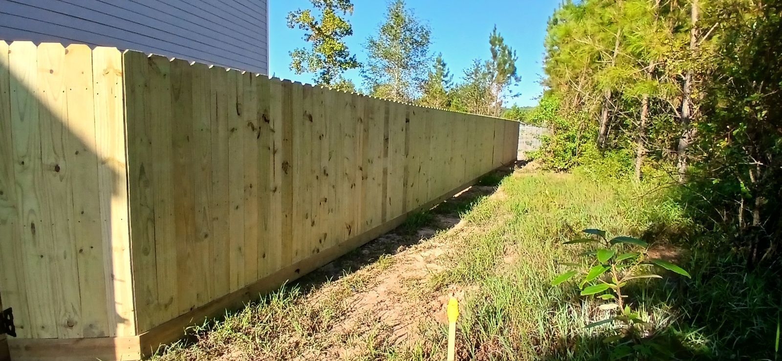 Wooden fence bordering a grassy area next to a building and trees under a clear blue sky.