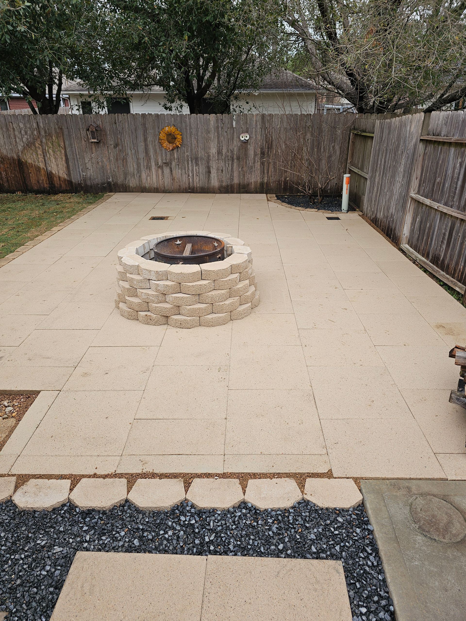 Backyard patio with fire pit, light beige pavers, and wooden fence. Black gravel border.