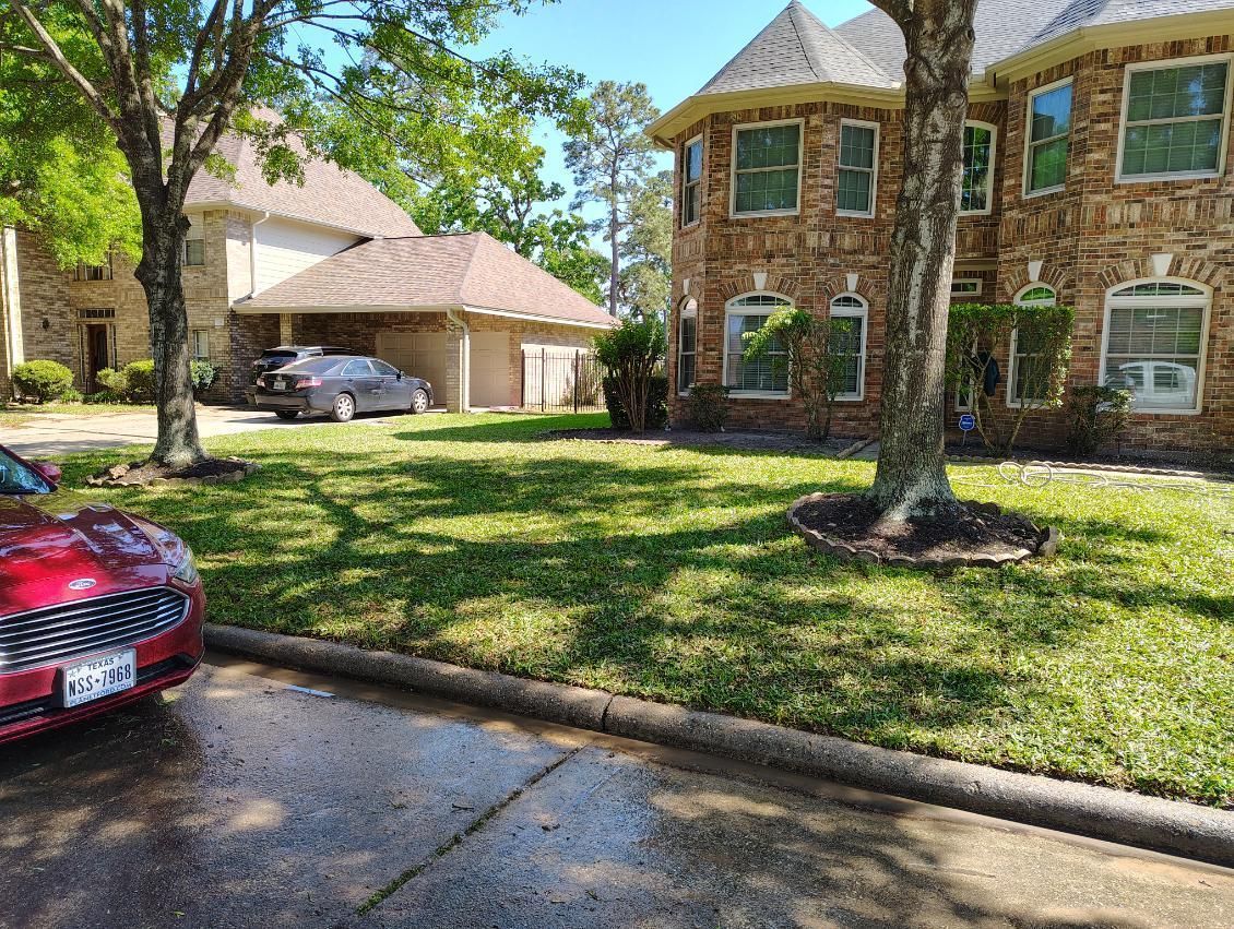 A red car parked in front of a large brick house with a green lawn and trees.