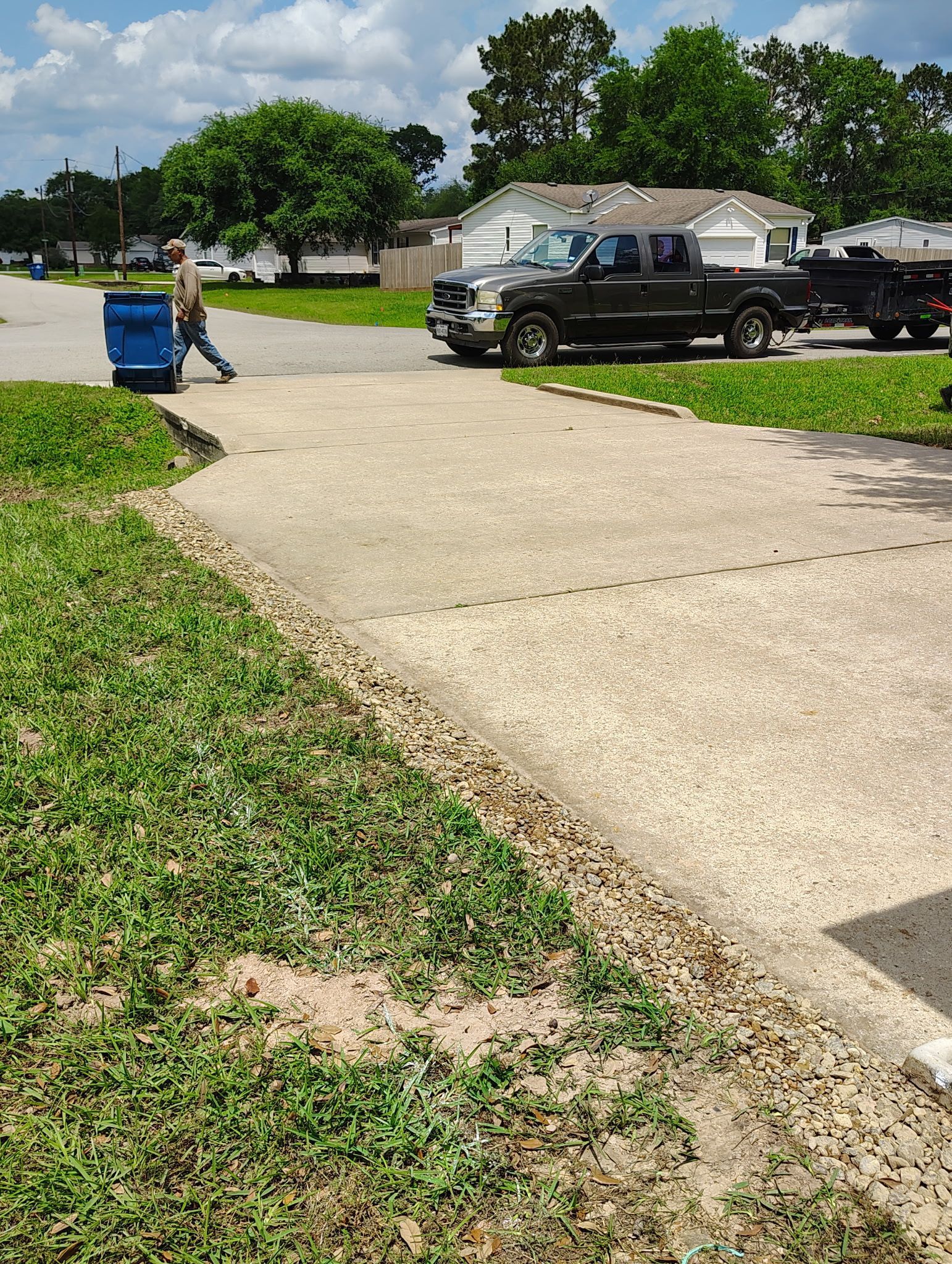 Man carrying blue bin on driveway near a truck. Green grass and houses in the background.