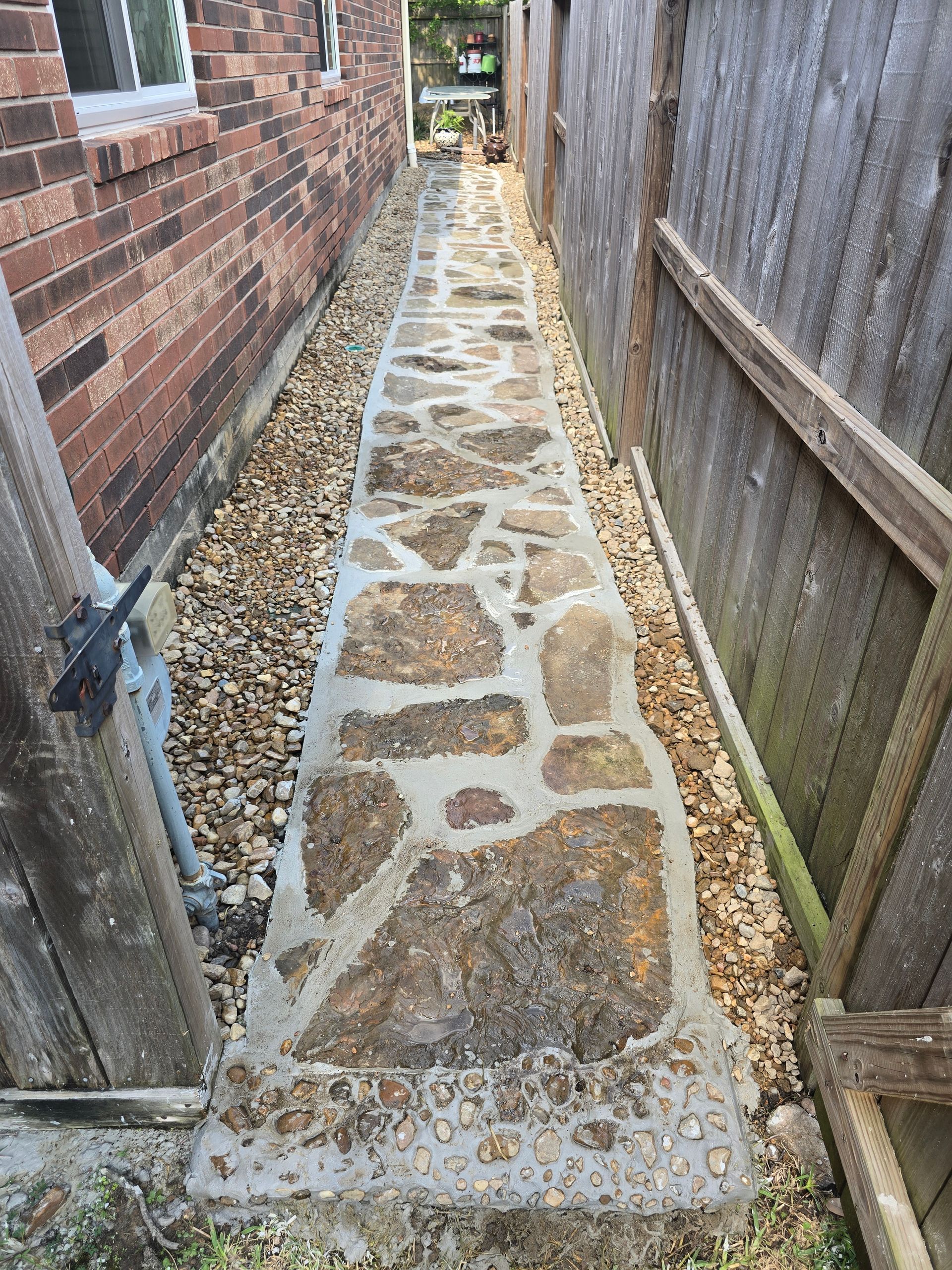 Narrow stone path between brick wall and wooden fence, with gravel bordering the path.