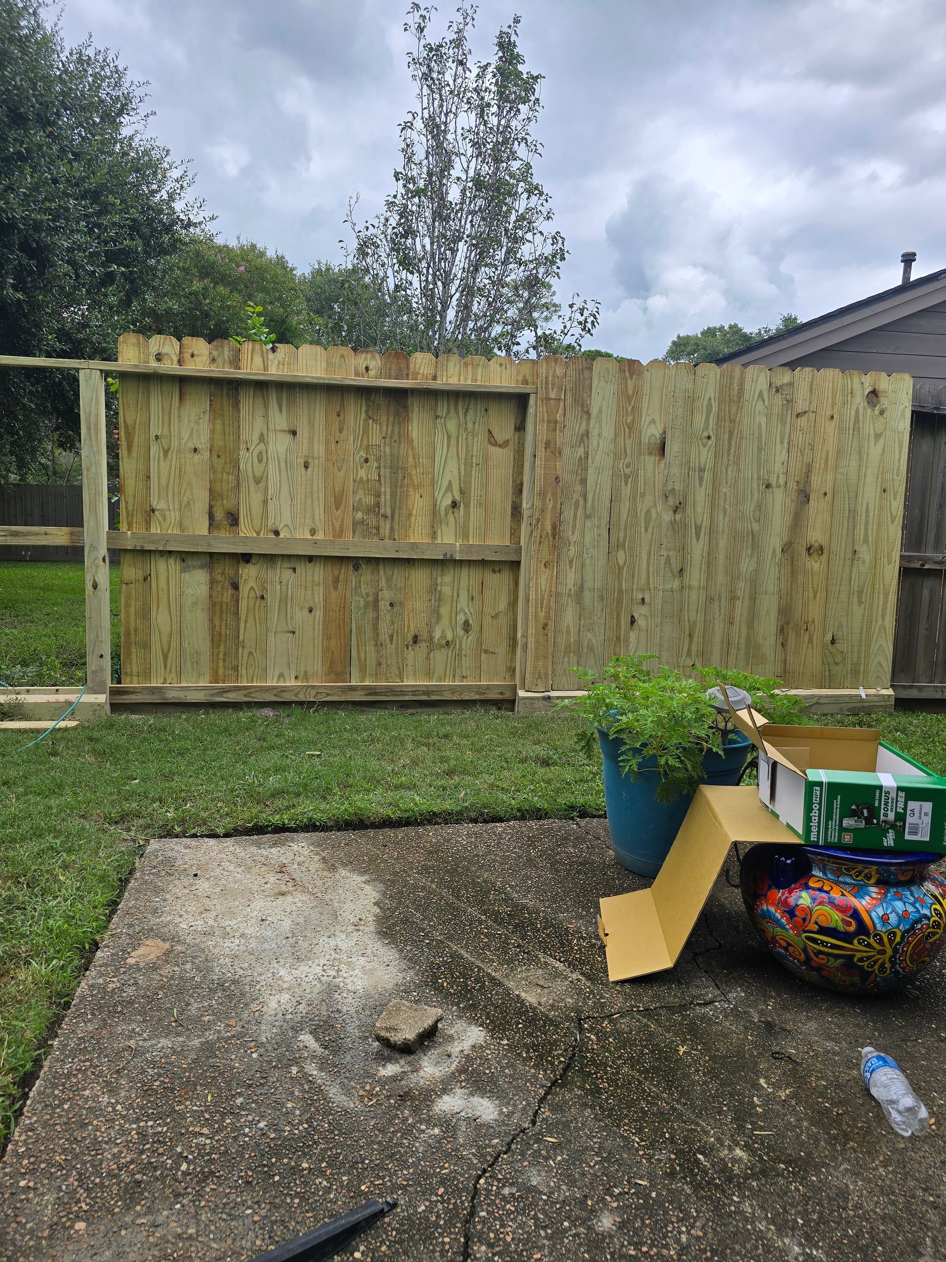Backyard with a wooden fence, blue planter, and concrete patio. Green grass and cloudy sky.