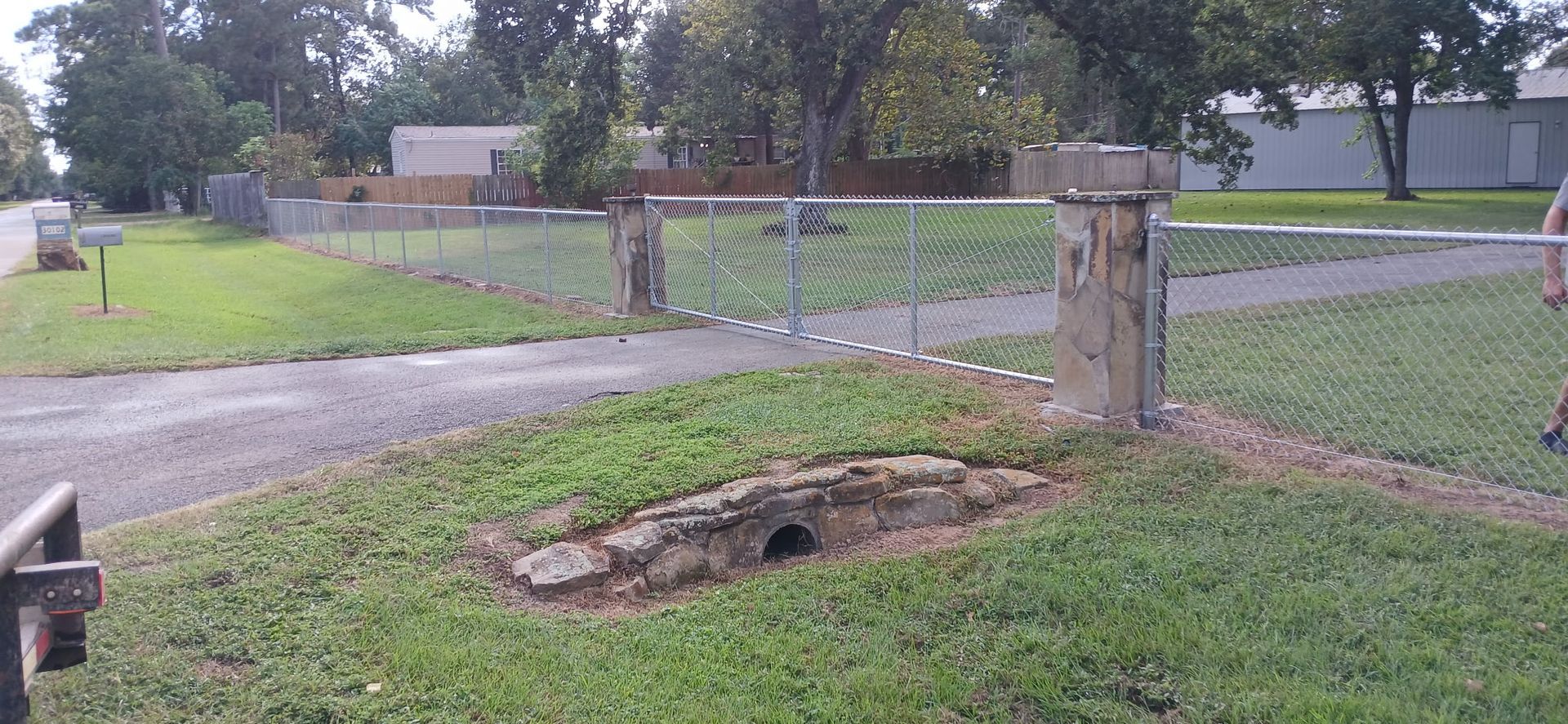A chain-link fence gate with stone pillars opens onto a driveway and grassy yard.
