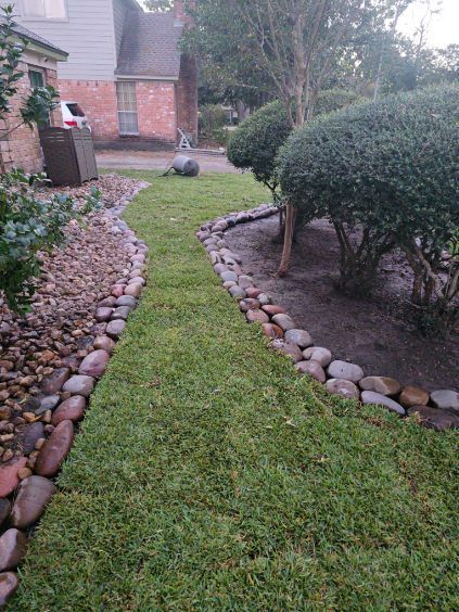 A grassy path bordered by rocks, leading towards a house with bushes and a statue.