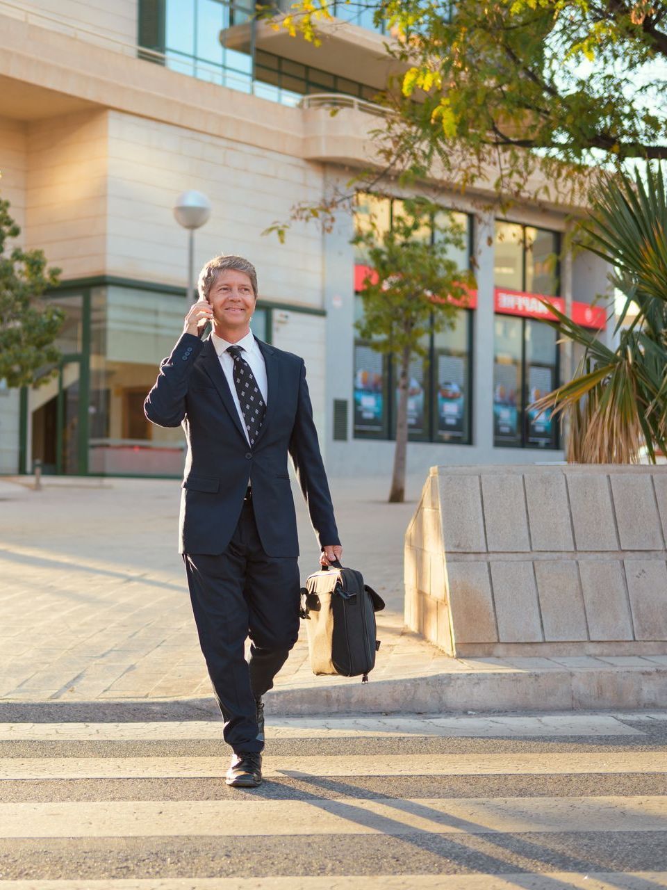 A man in a suit and tie is crossing the street while talking on a cell phone