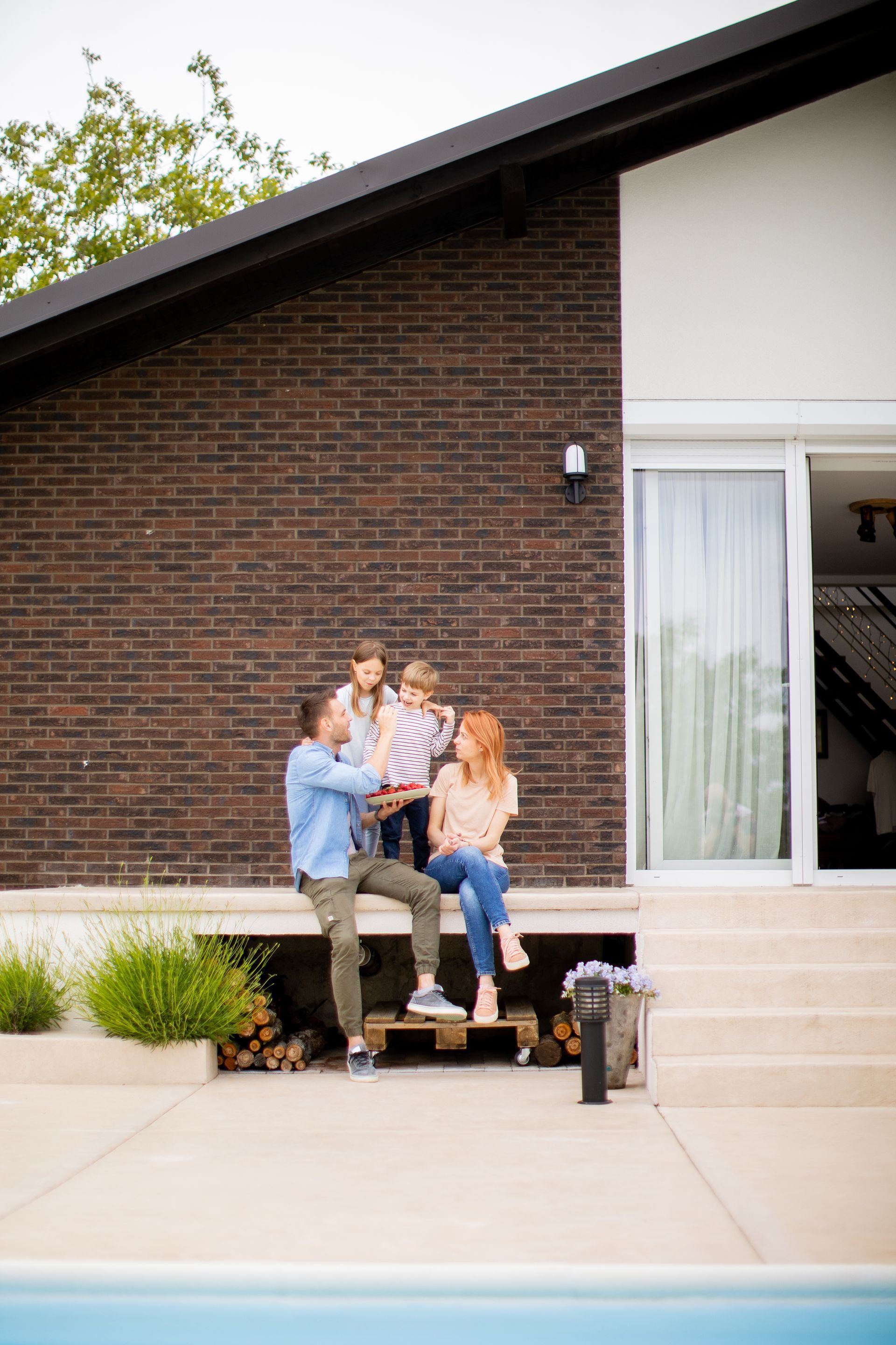 A family is sitting on the porch of their house next to a swimming pool.