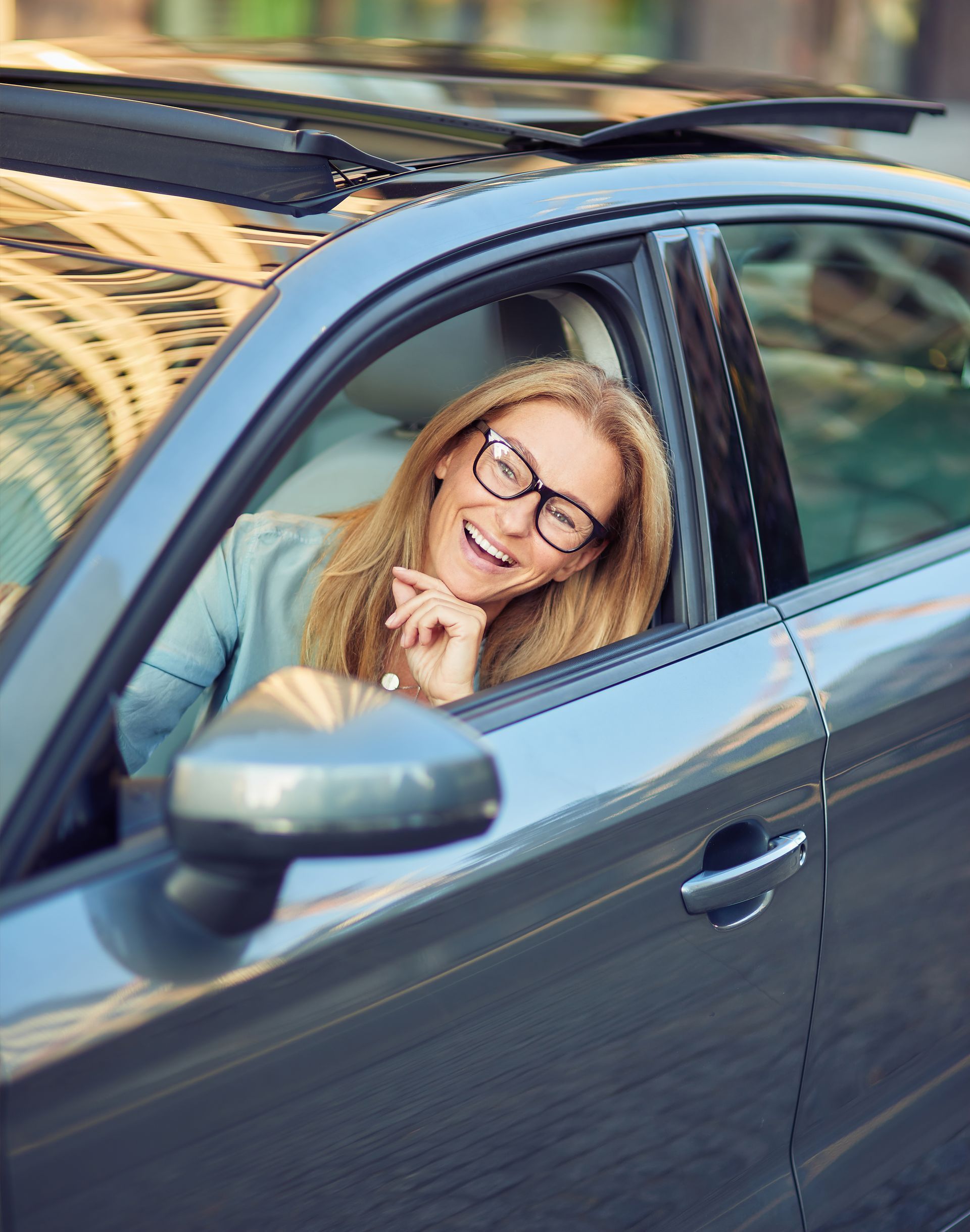 A woman wearing glasses is sitting in a car and smiling.