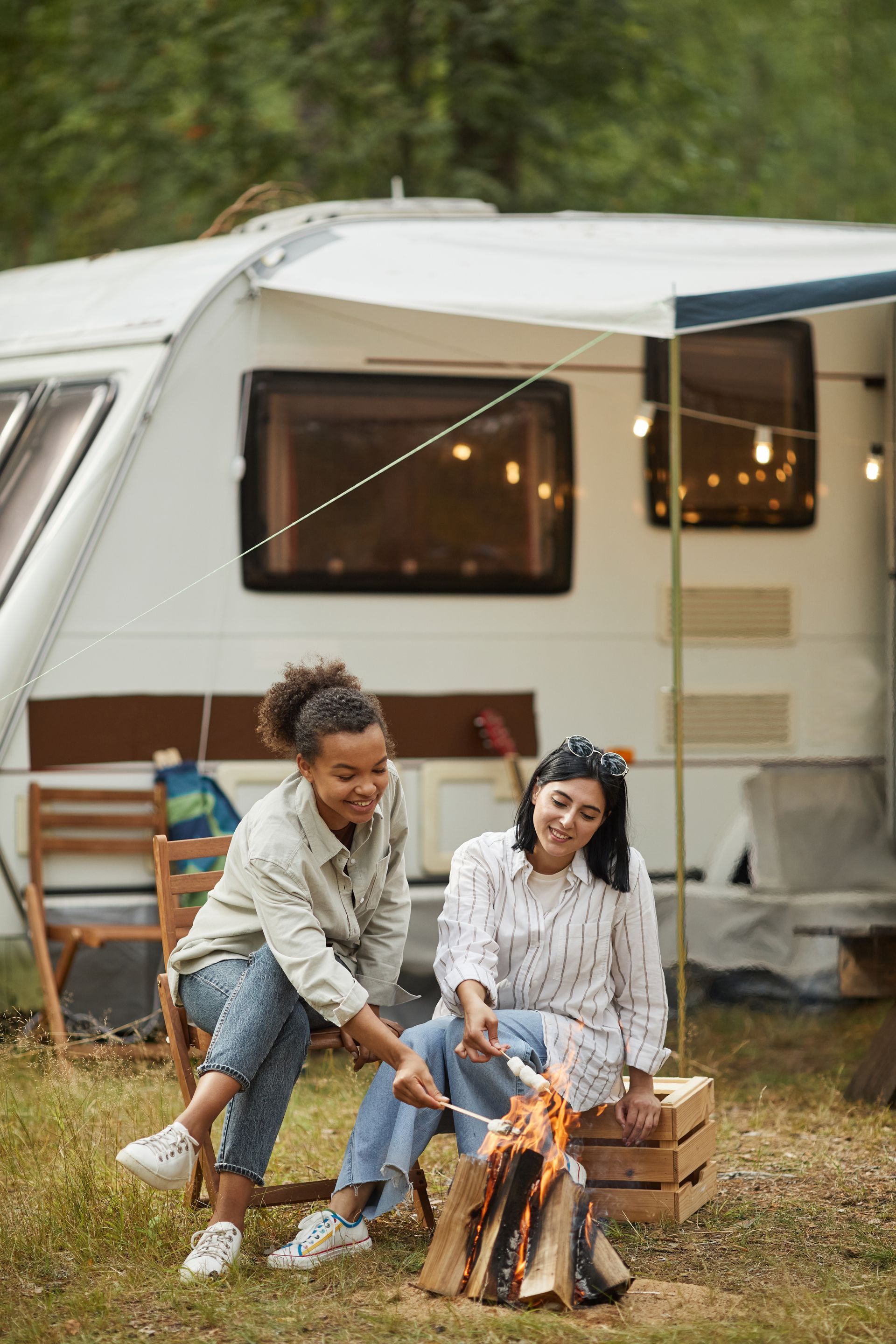 Two women are roasting marshmallows over a campfire in front of a trailer.