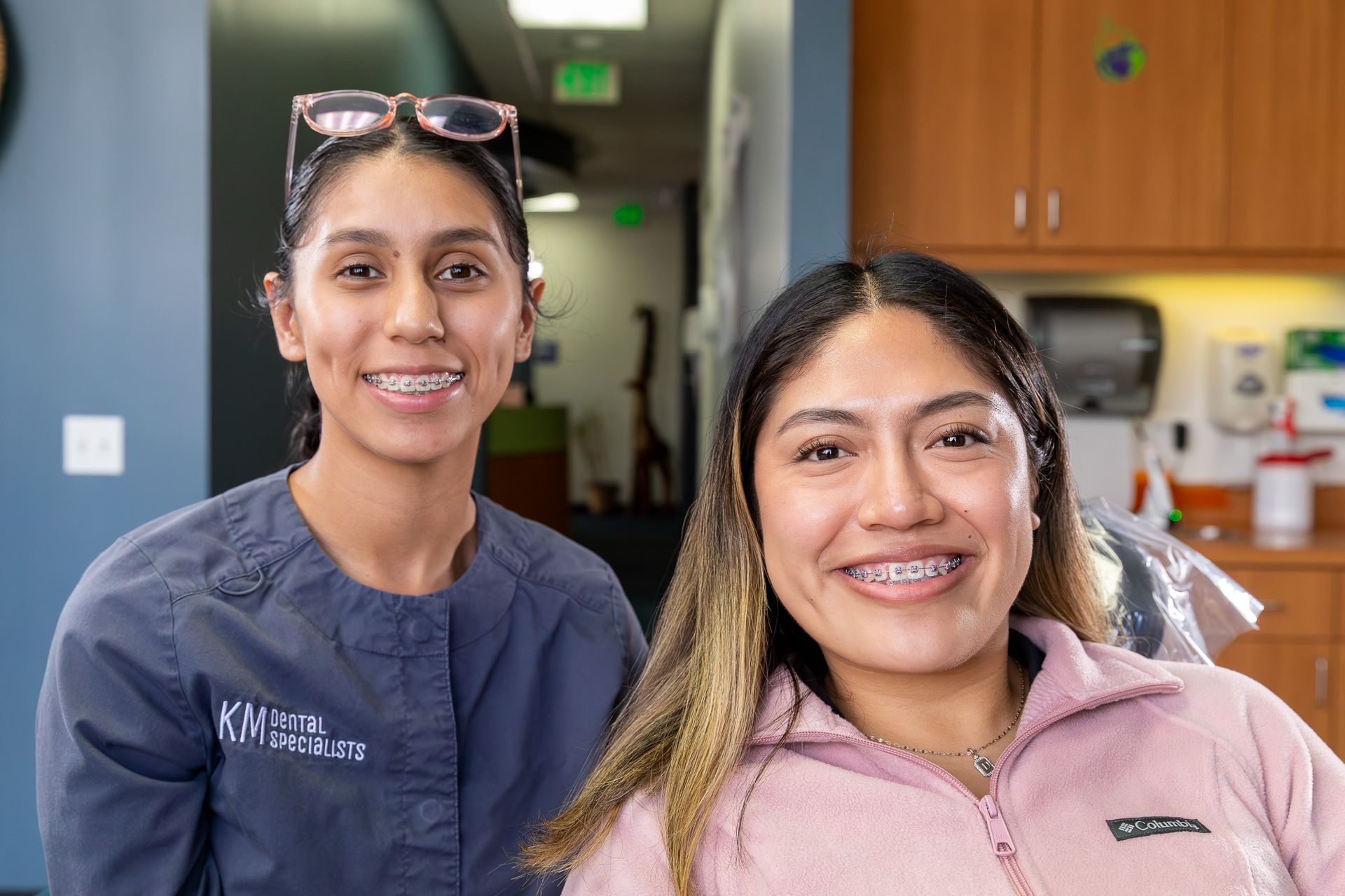 Two smiling coworkers in a kitchen or break room, one in navy scrubs and one in a pink jacket.
