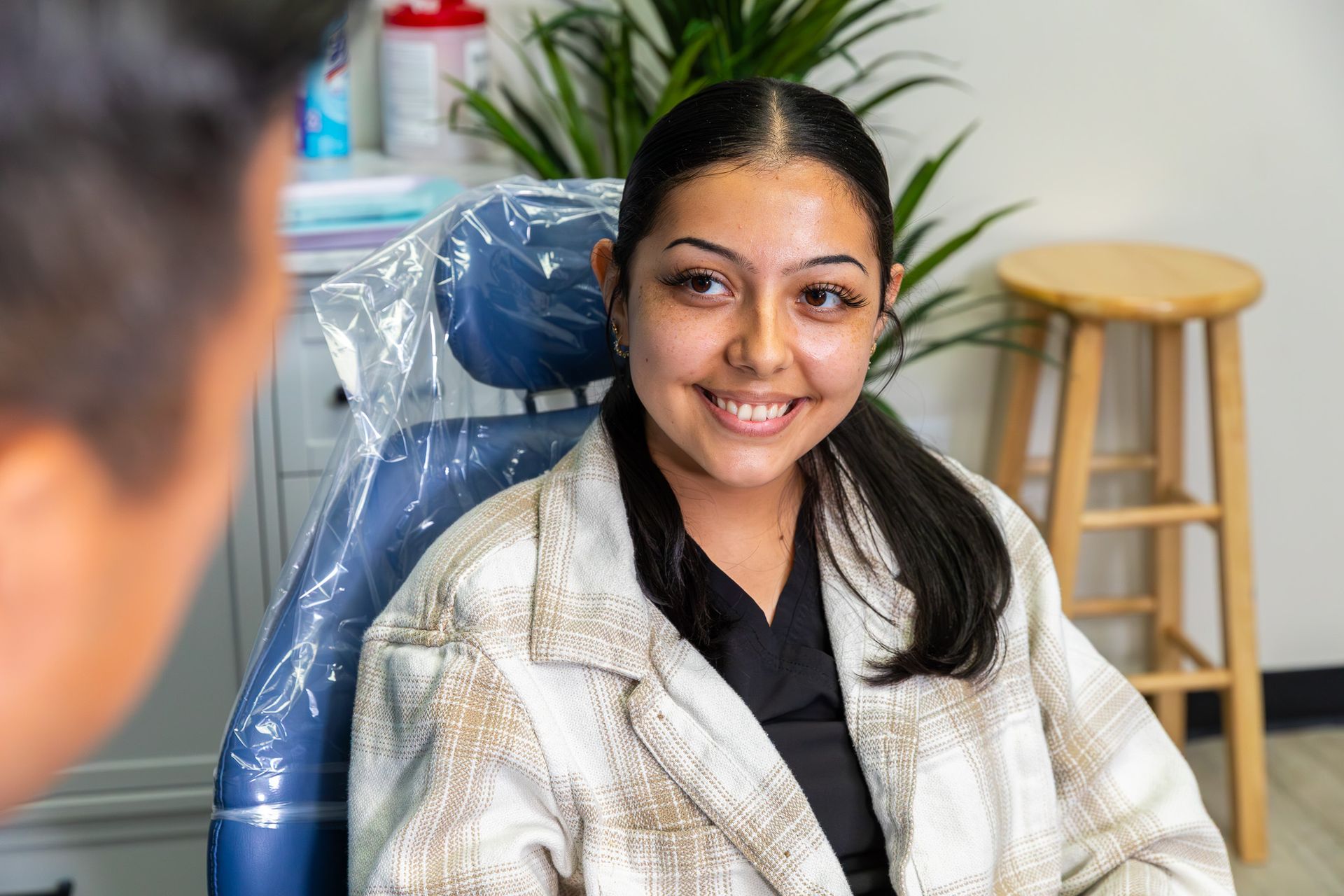 Woman smiling in a dental chair, facing a person. She wears a plaid jacket and black top. Clinic setting.