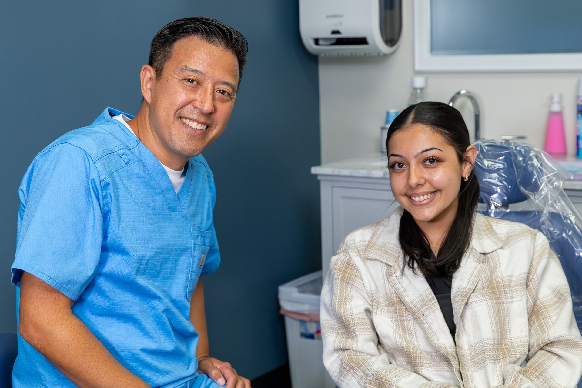 Dentist in blue scrubs smiles next to a smiling patient seated in a dental chair.