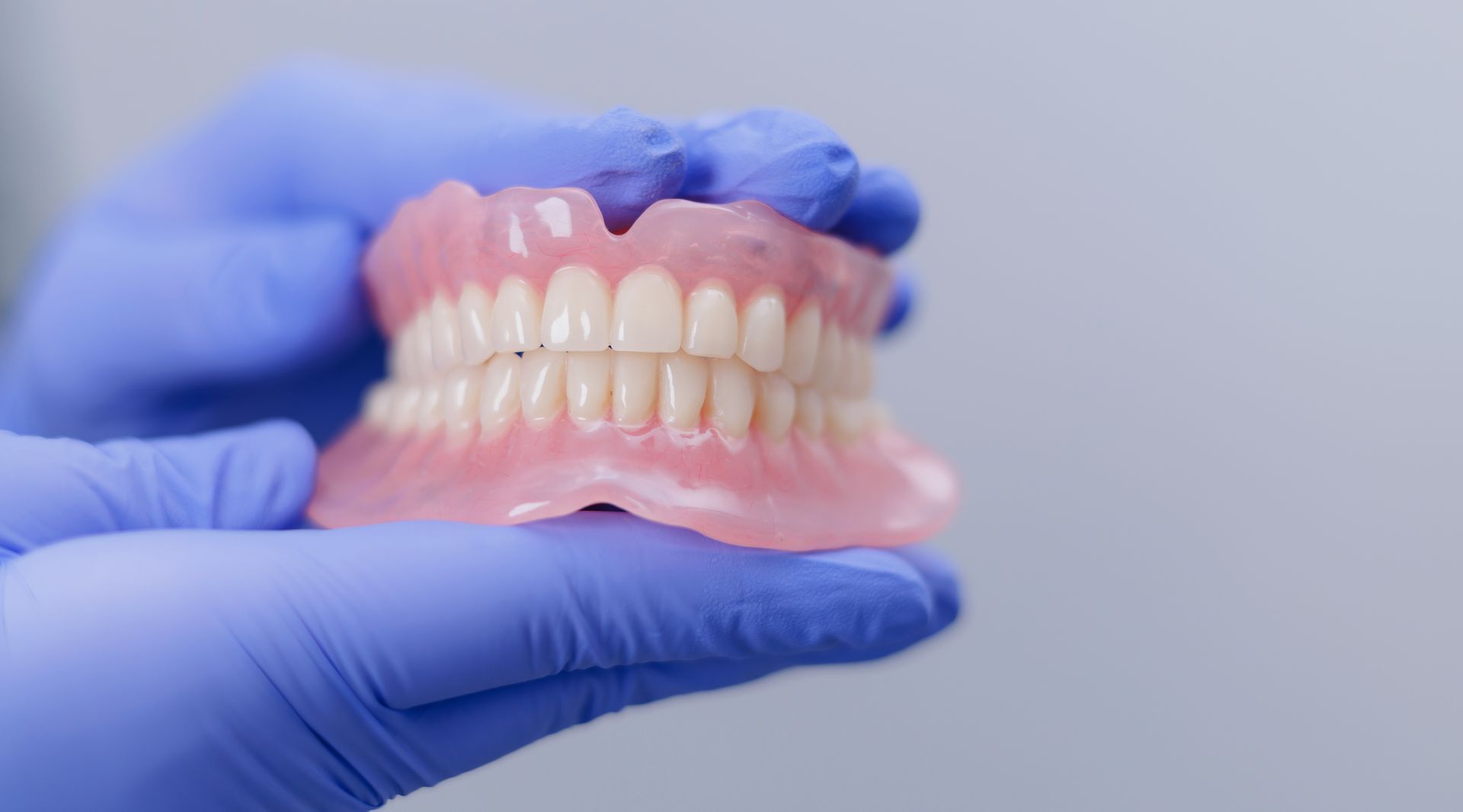 Gloved hands holding a pink and white dentures model against a plain background