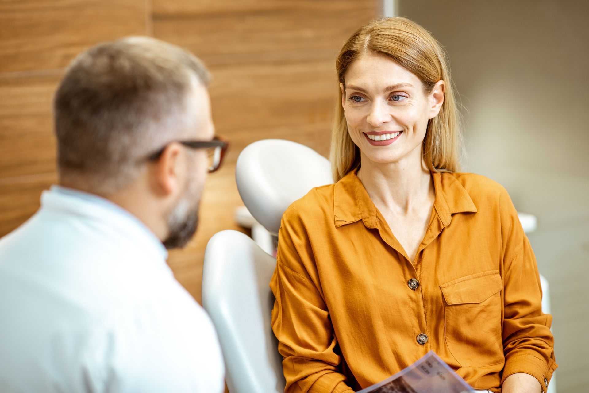 Two people talk in a dental office; one smiles while seated in an orange shirt.