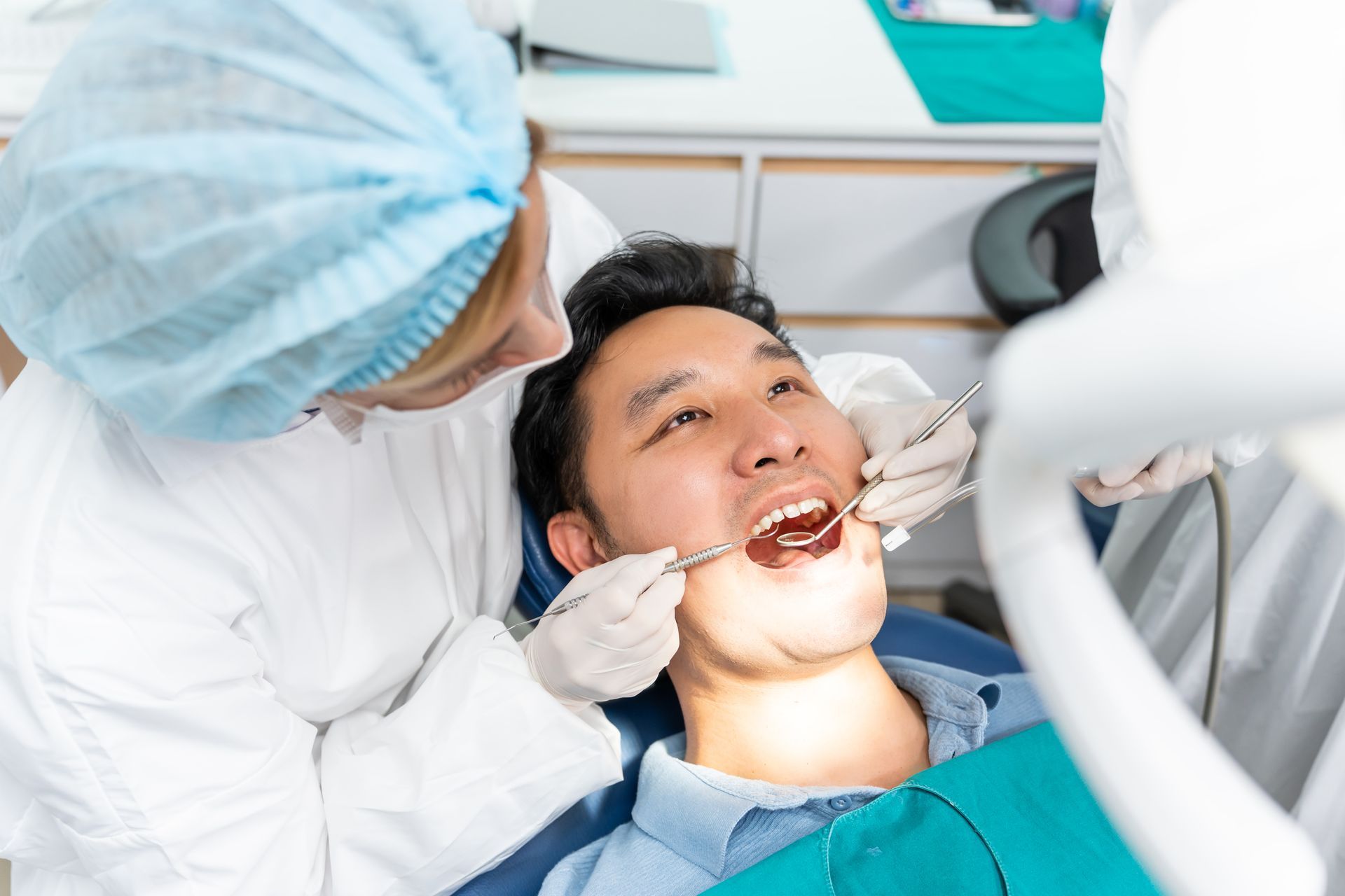 A dentist in a blue cap and white coat examines a patient’s teeth using a dental mirror and probe in a clinic.