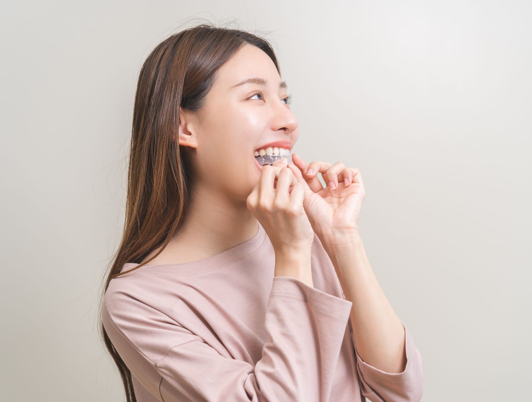 Woman smiling, inserting clear aligner in mouth, light pink shirt, neutral background.
