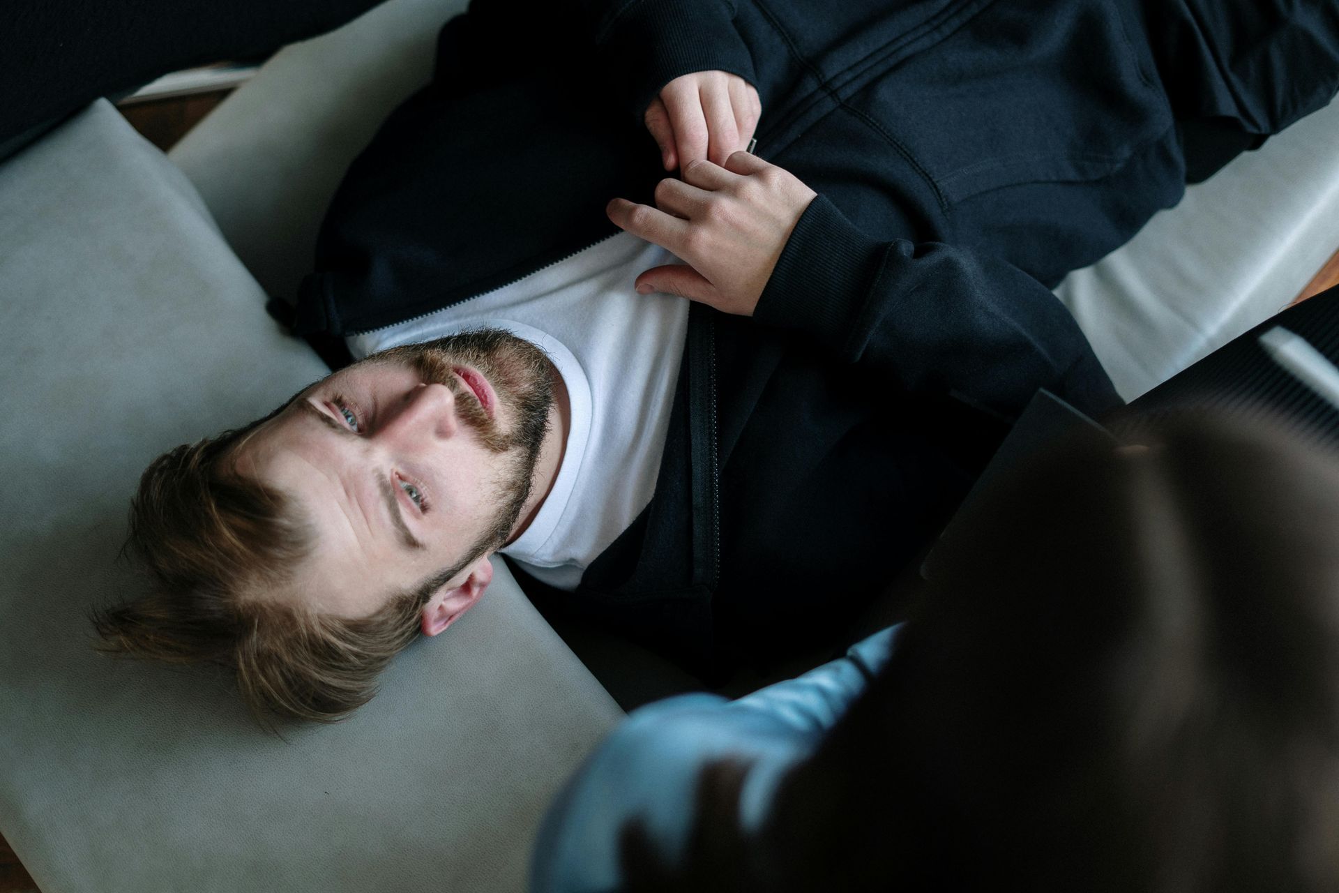 Man lying on a gray couch, wearing a black jacket and white shirt. Eyes closed, relaxed expression.