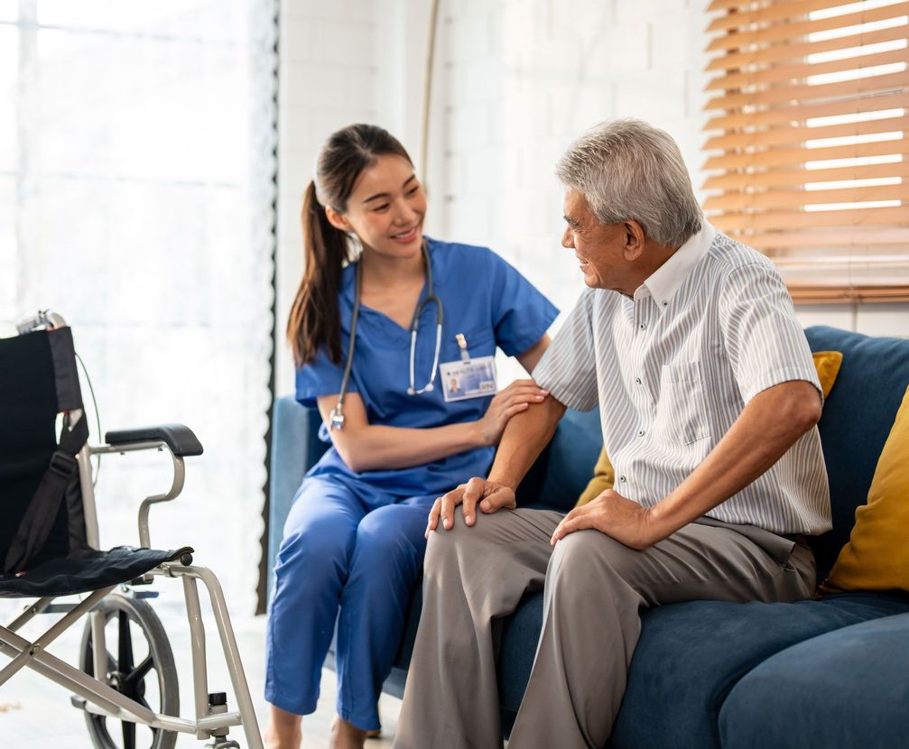 A young male caregiver in blue scrubs assists an elderly man with a cane in a home kitchen.